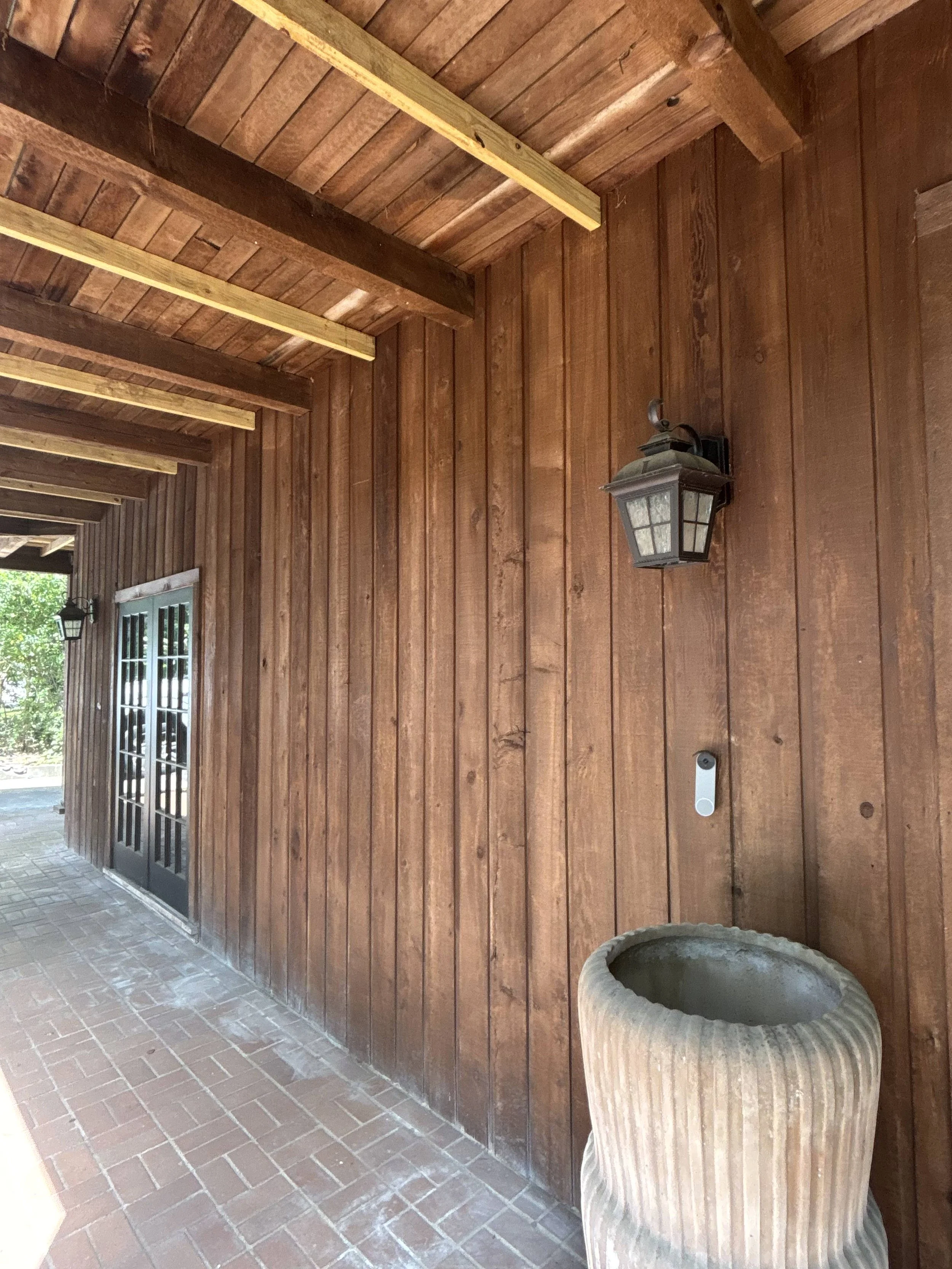 Exterior view of a wooden porch with a lantern light, a ceramic planter, and a glass-pained door with black framing.