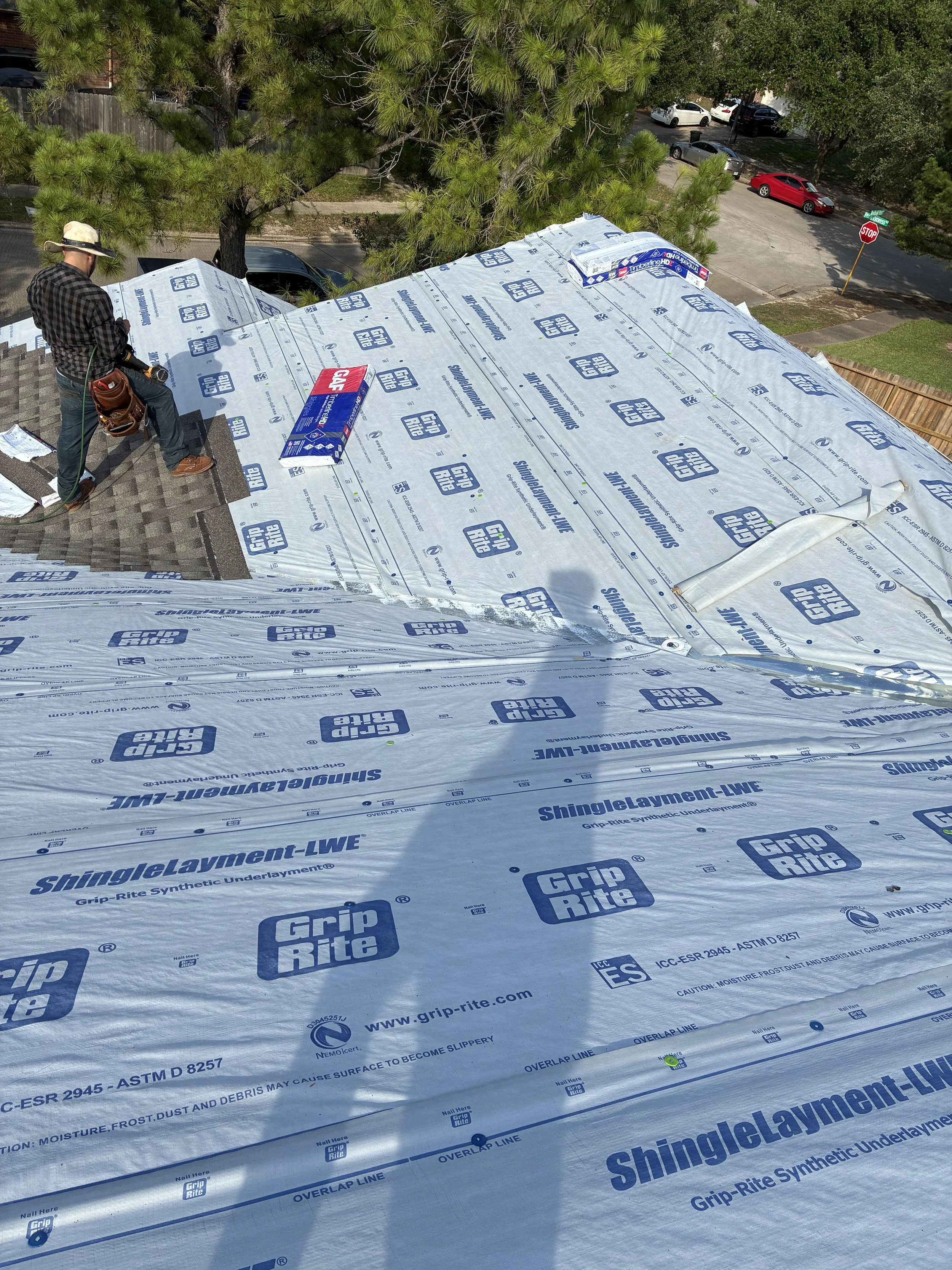 A person working on roofing a house, using construction materials labeled 'Grip Rite' and 'ShingleLayment-LWE' under a tree with parked cars and a stop sign in the background.