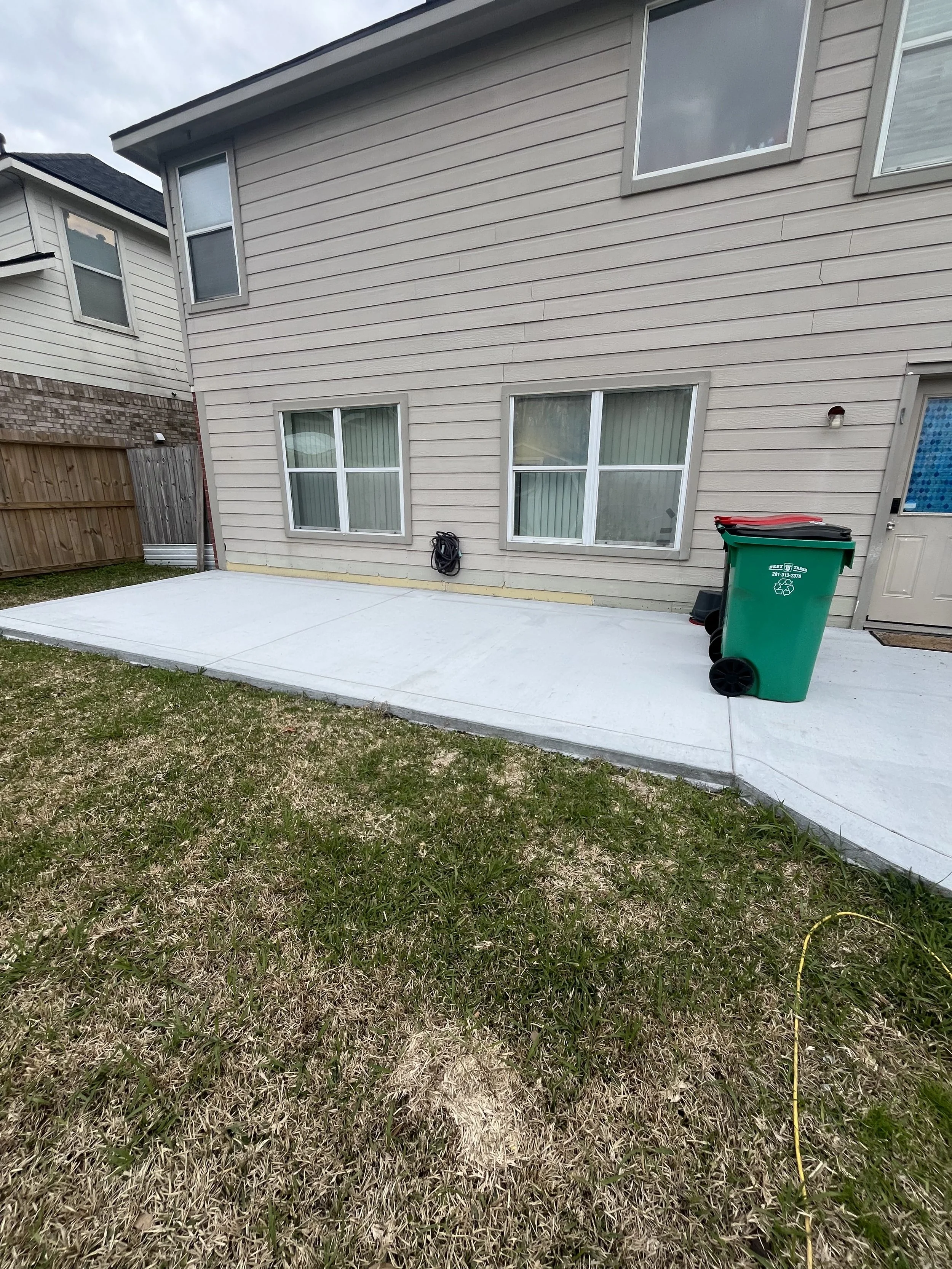 Backyard with a concrete patio, beige siding house with windows, a green trash bin, and a garden hose.