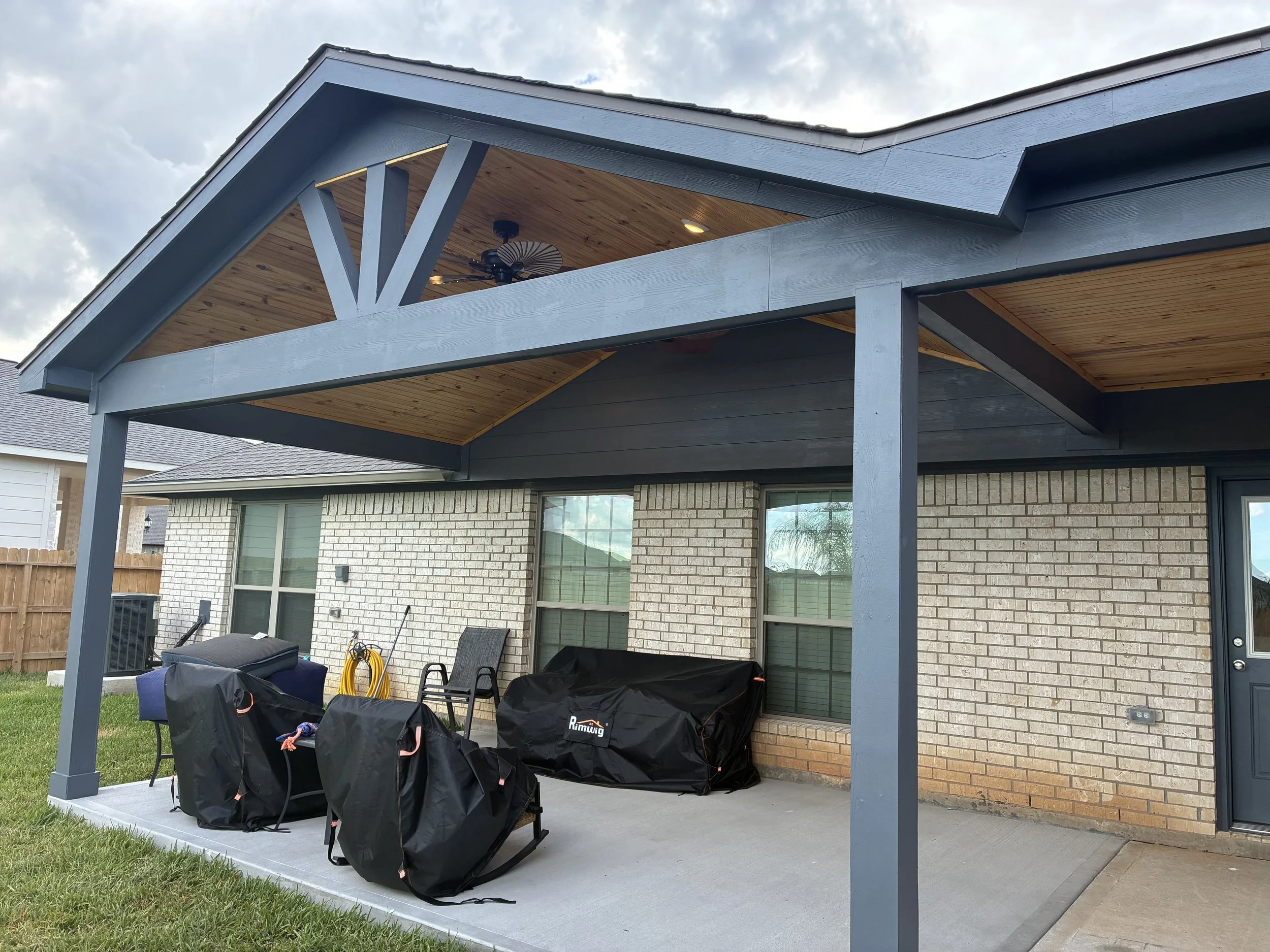 Back patio of a house with covered porch, BBQ grills covered with black covers, chairs, garden hose, and a yellow extension cord, with two windows and a door on brick wall, and a wooden fence in the background.