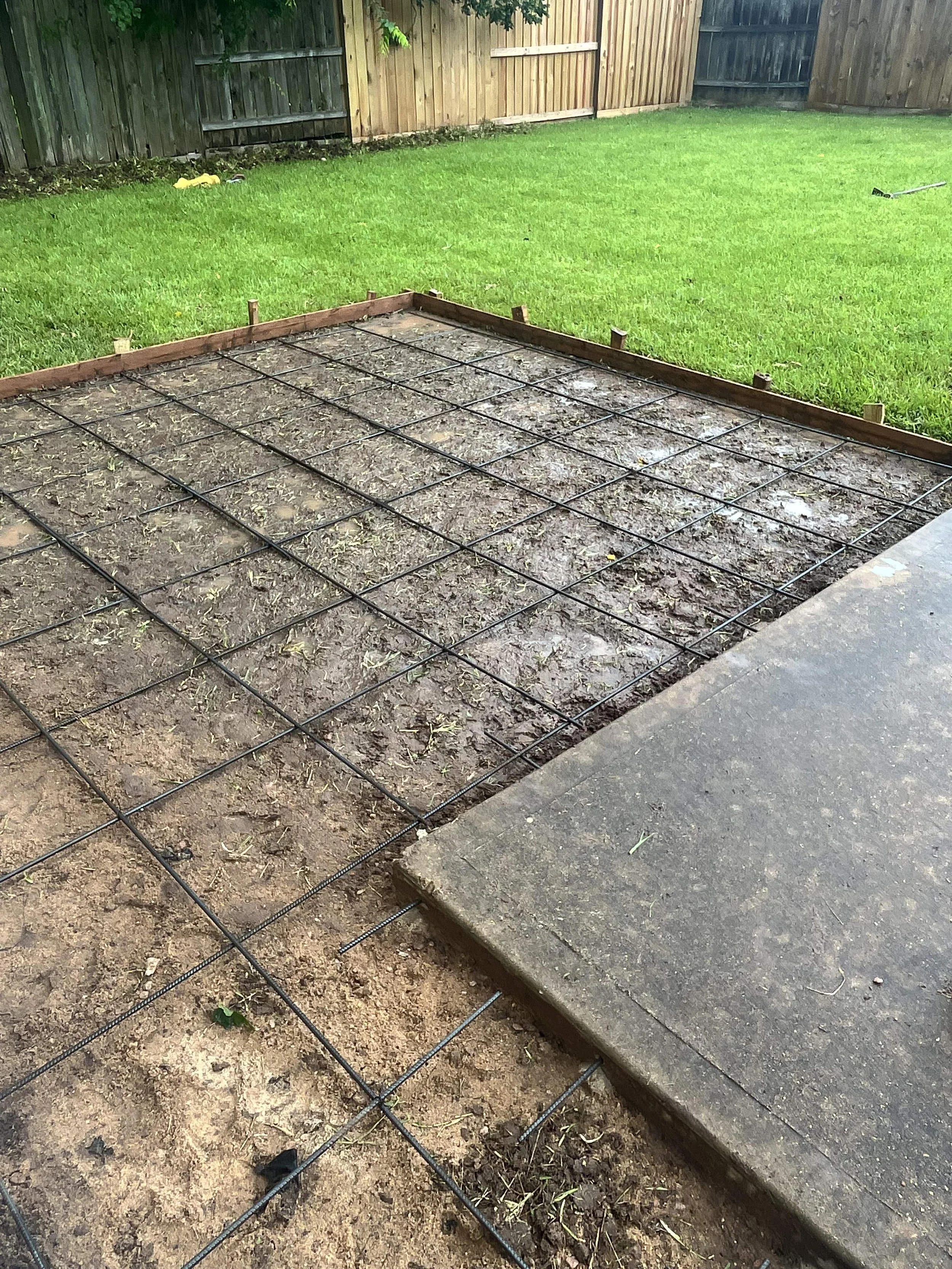A backyard with a section of ground prepared for concrete, with rebar grid reinforcement in place, surrounded by grass and wooden fencing.