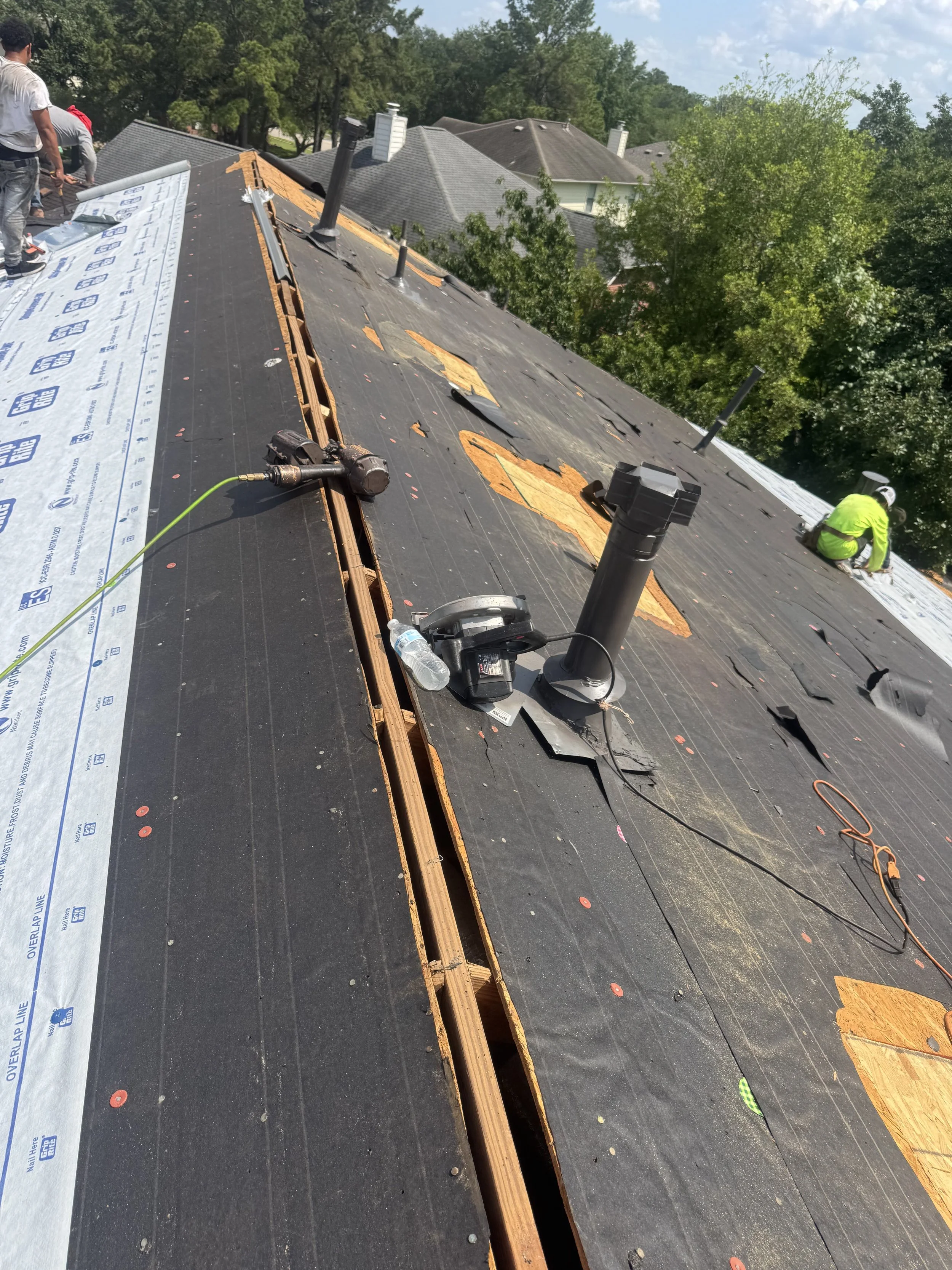 Roof under construction with workers installing new roofing material. The roof has black underlayment and is partially covered with protective tarp, with visible vents and tools.
