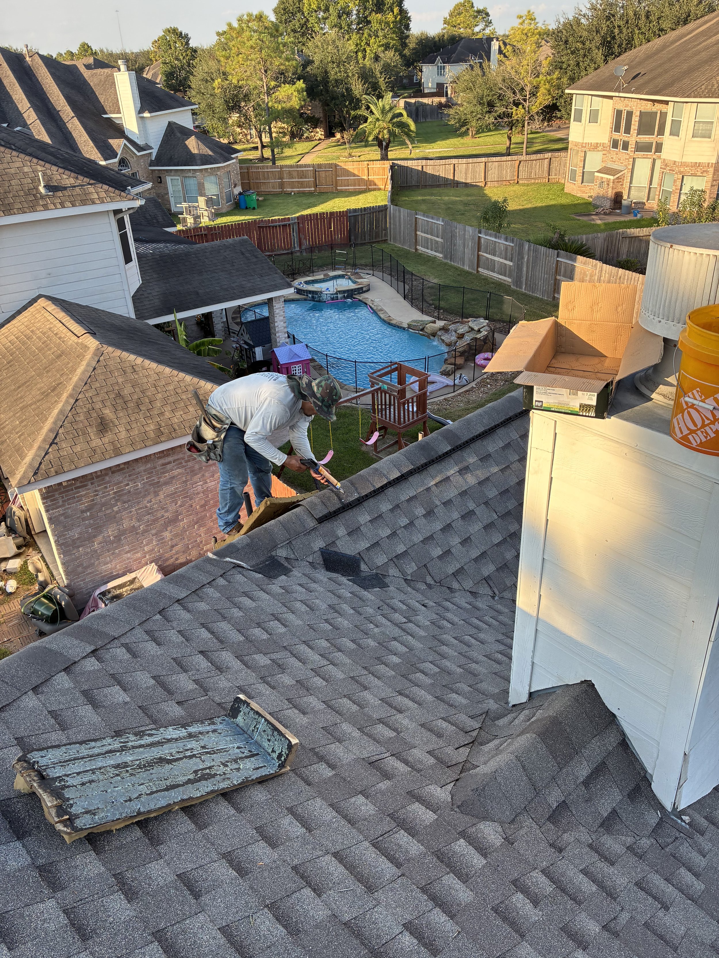 A person working on a roof with roofing tools, overlooking a backyard with a pool and neighboring houses.