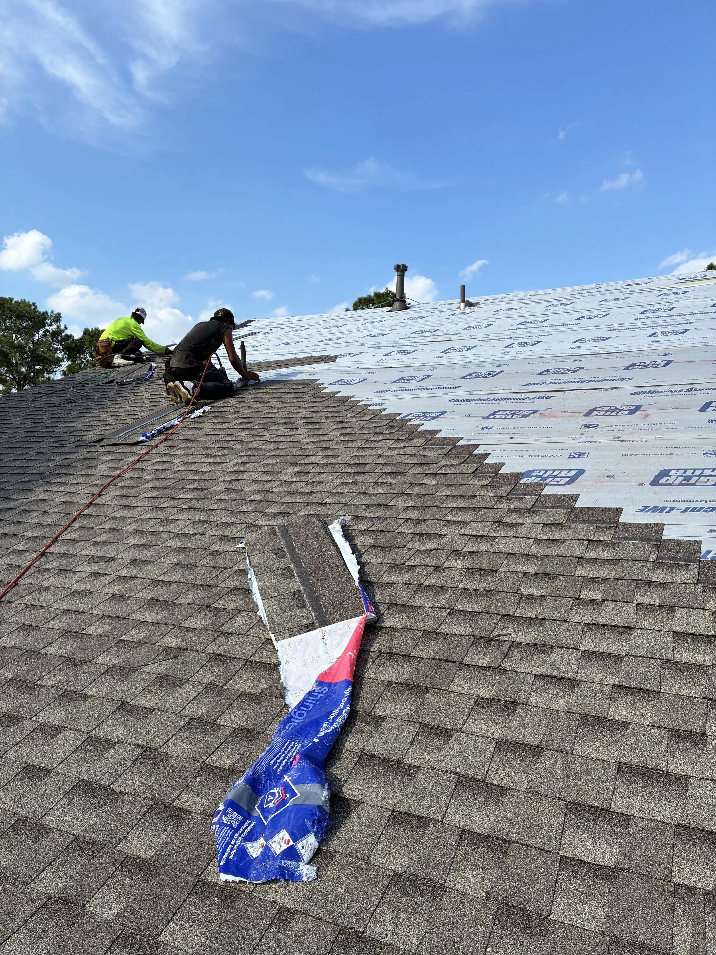 Construction workers installing new roofing shingles on a house under a blue sky.