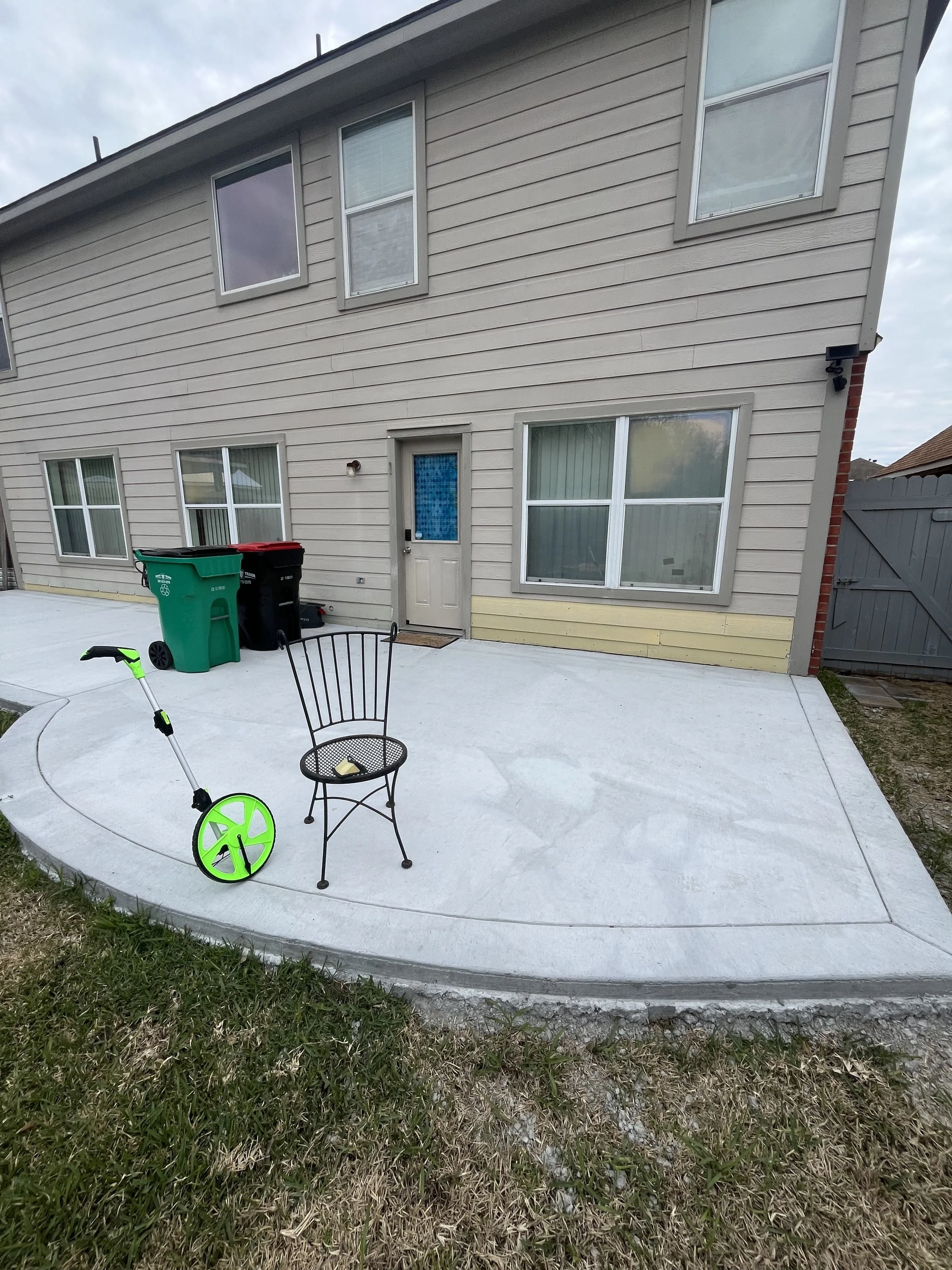 Backyard with a concrete patio, two trash bins, a black and green scooter, a black metal chair, and the back of a beige house with multiple windows and a door.