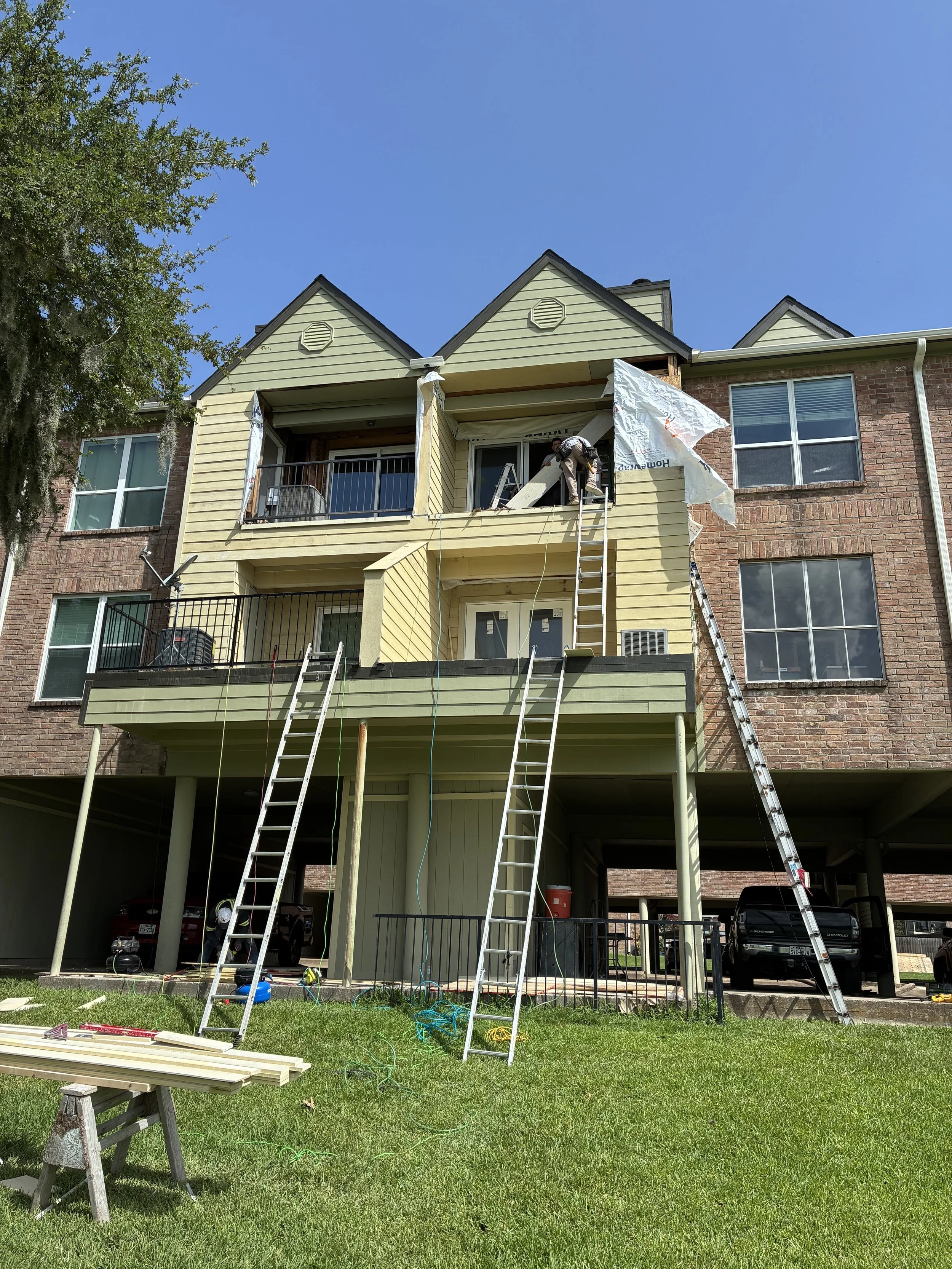 Construction workers installing siding on a multi-story residential building, with ladders, tools, and building materials nearby.