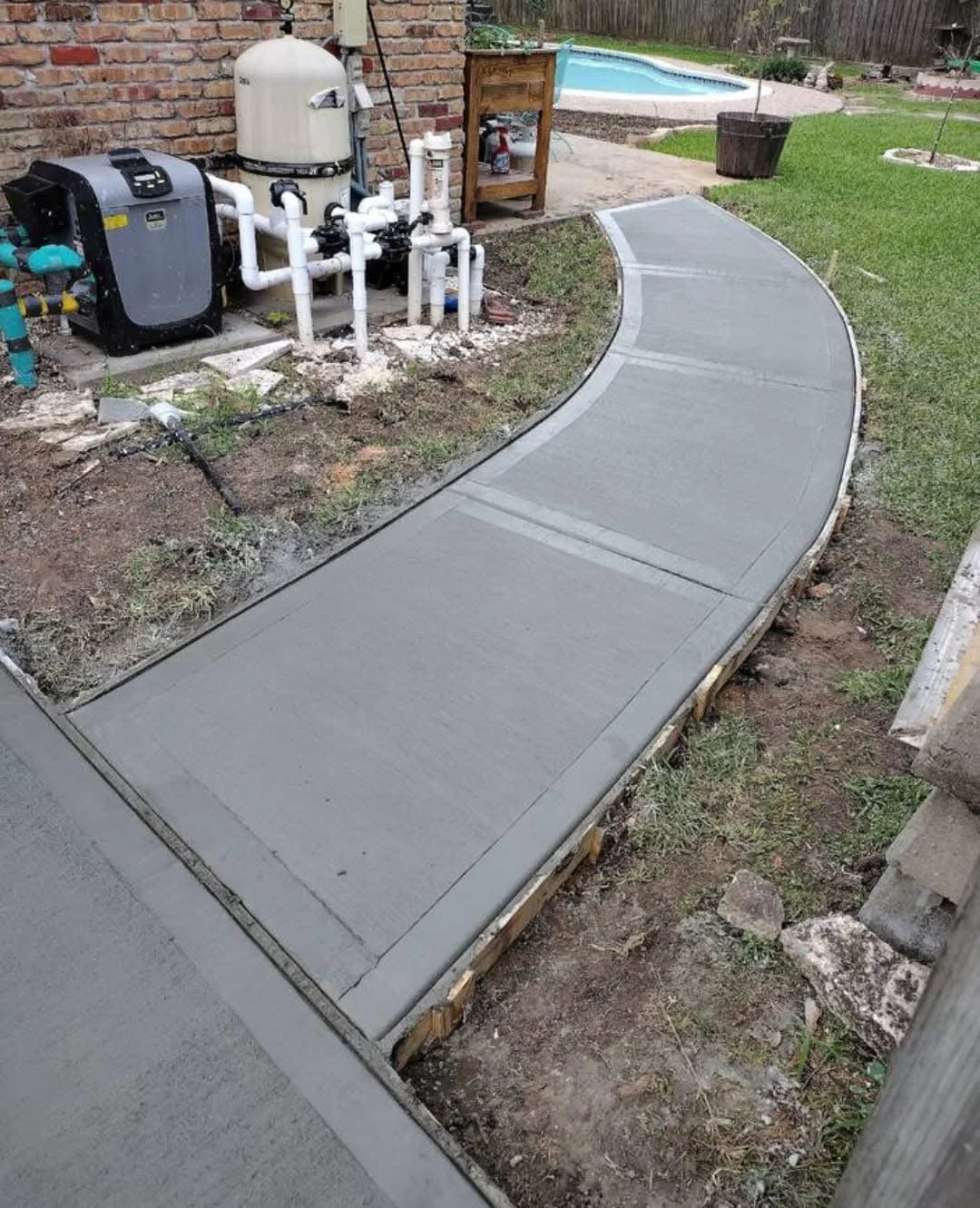 Newly poured concrete sidewalk with a curved design in a backyard, alongside a brick wall with pool equipment and piping on the side, and a small in-ground pool in the background.