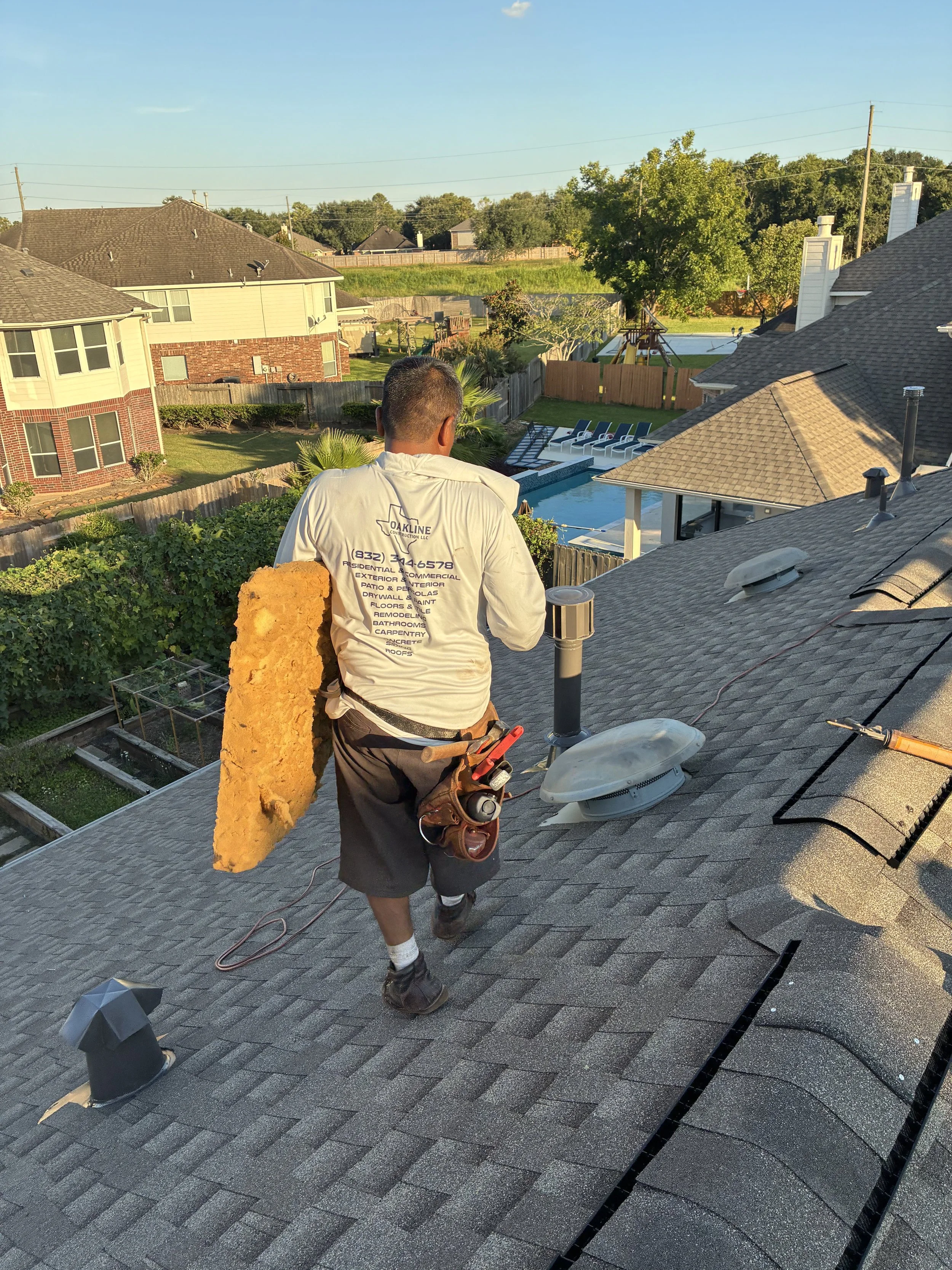 A worker walking on a residential roof holding a large piece of roofing material. The worker is wearing a white shirt with company details, shorts, and tool belt. The background shows neighboring houses, a backyard pool, and a bright, clear sky.