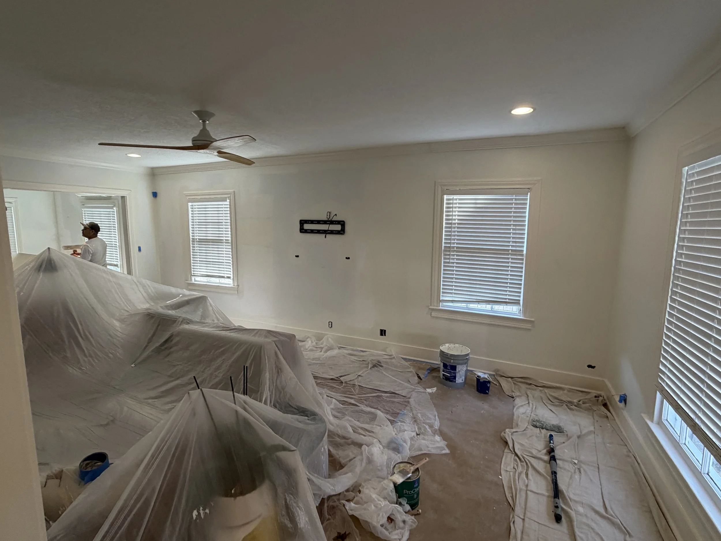 Living room under renovation with covered furniture, painter's supplies, and a worker in the background.