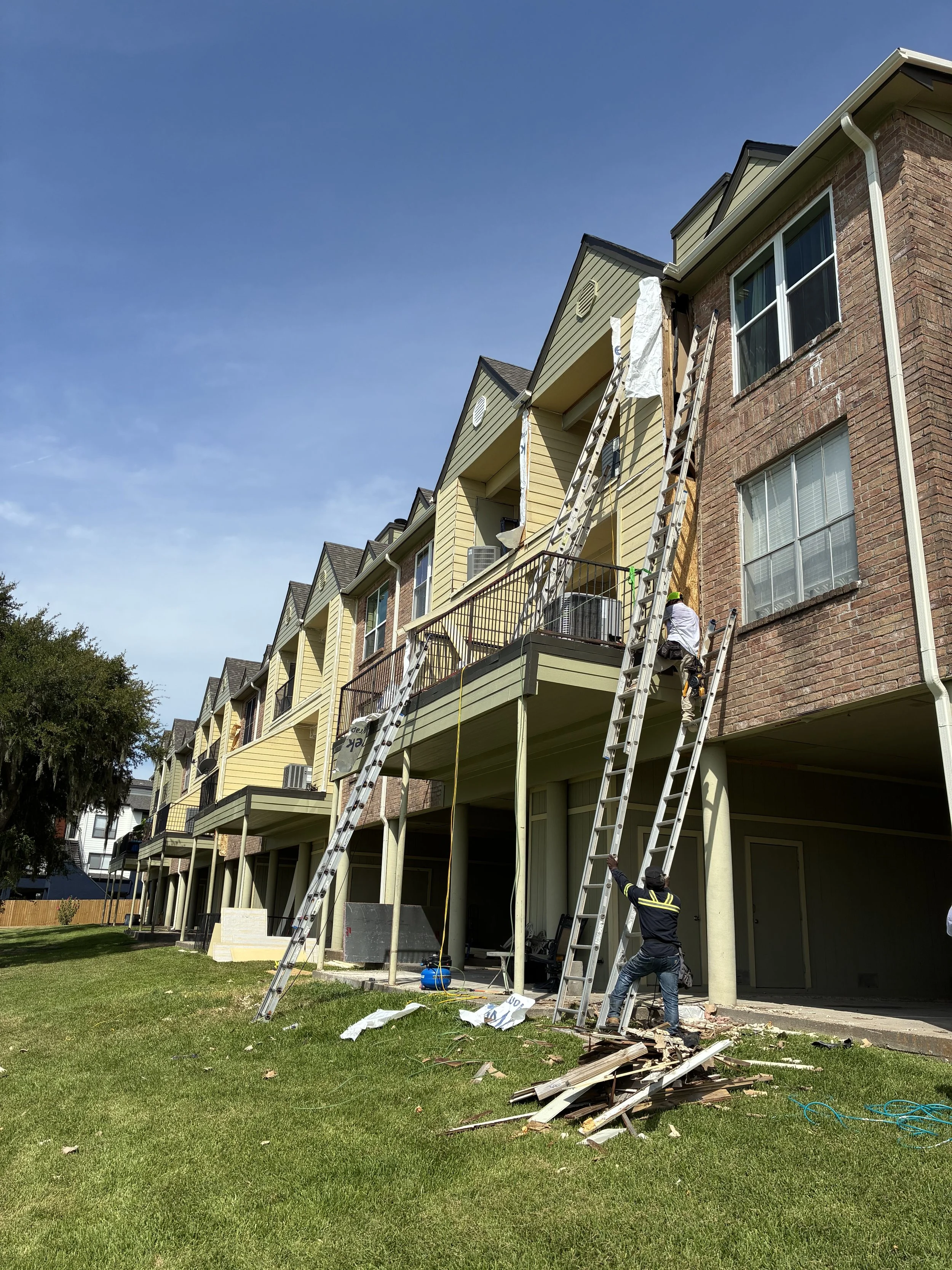 Construction workers repairing the side of a multi-story residential building with ladders and debris on the ground.
