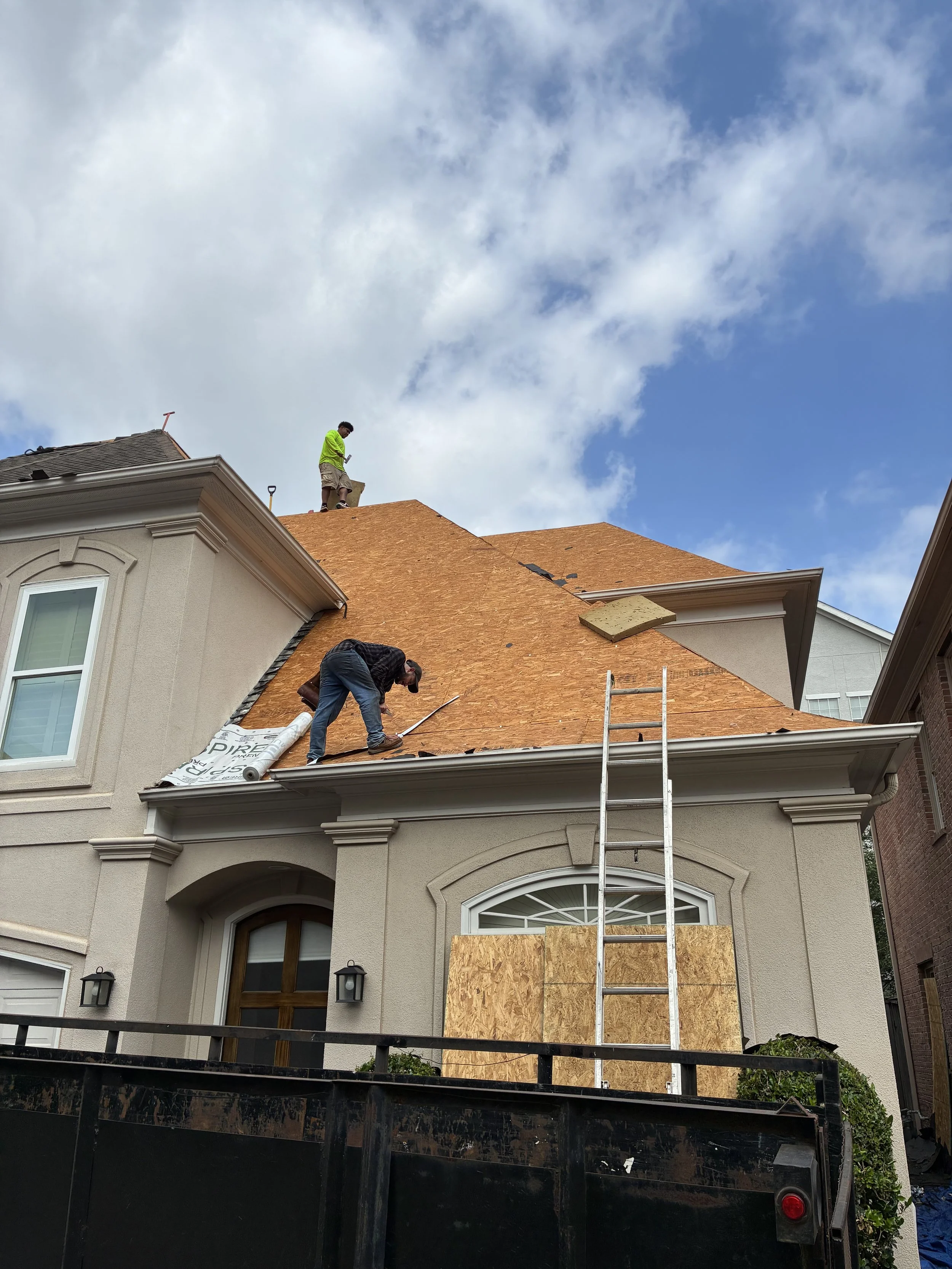 Two workers on the roof of a house installing new plywood sheathing, with a ladder leaning against the front of the house. The house has white siding and arched windows, and the sky is partly cloudy.