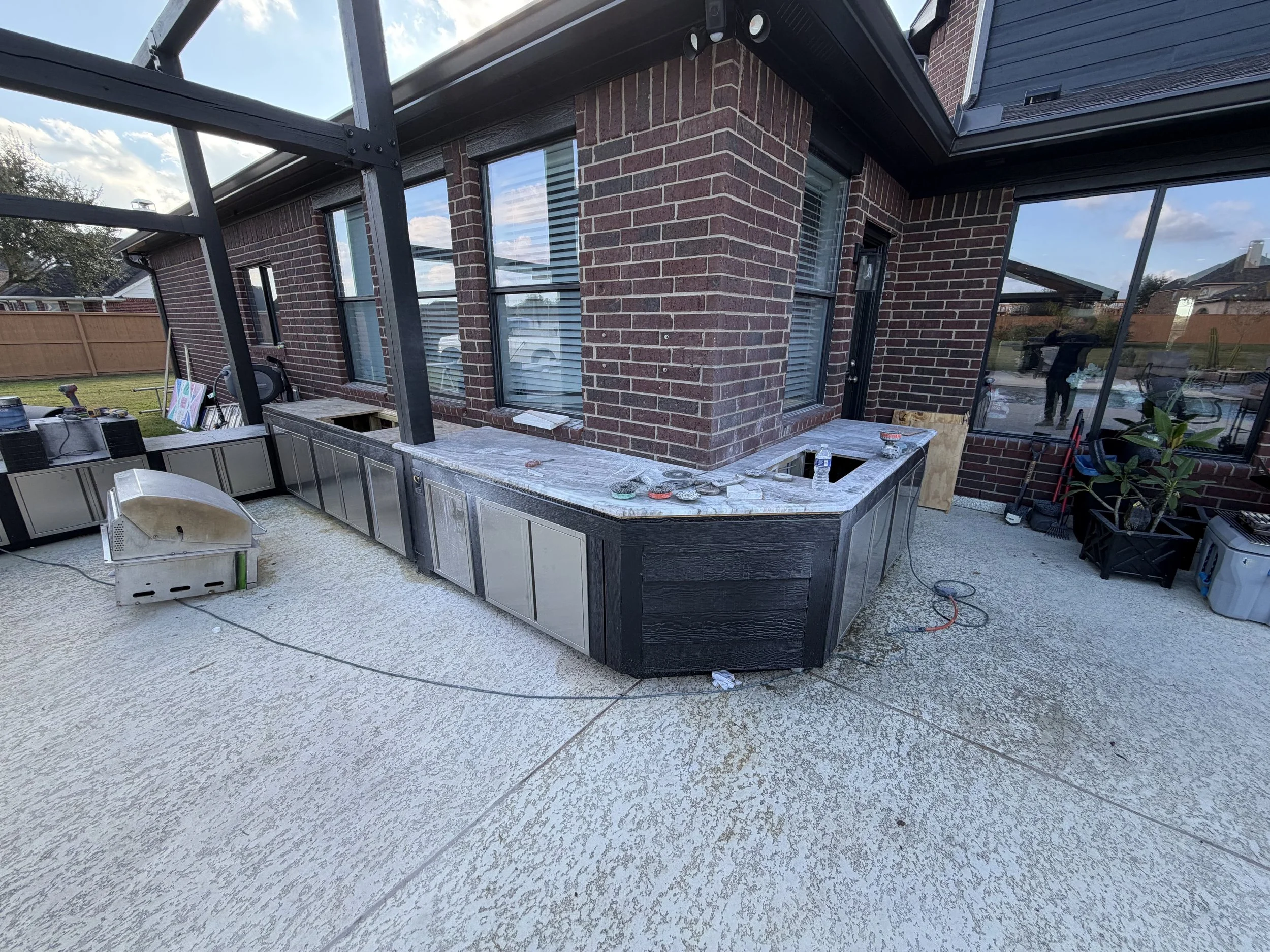 Backyard patio under construction, with a brick house, large glass windows, and an outdoor kitchen area with countertops, cabinets, and tools.