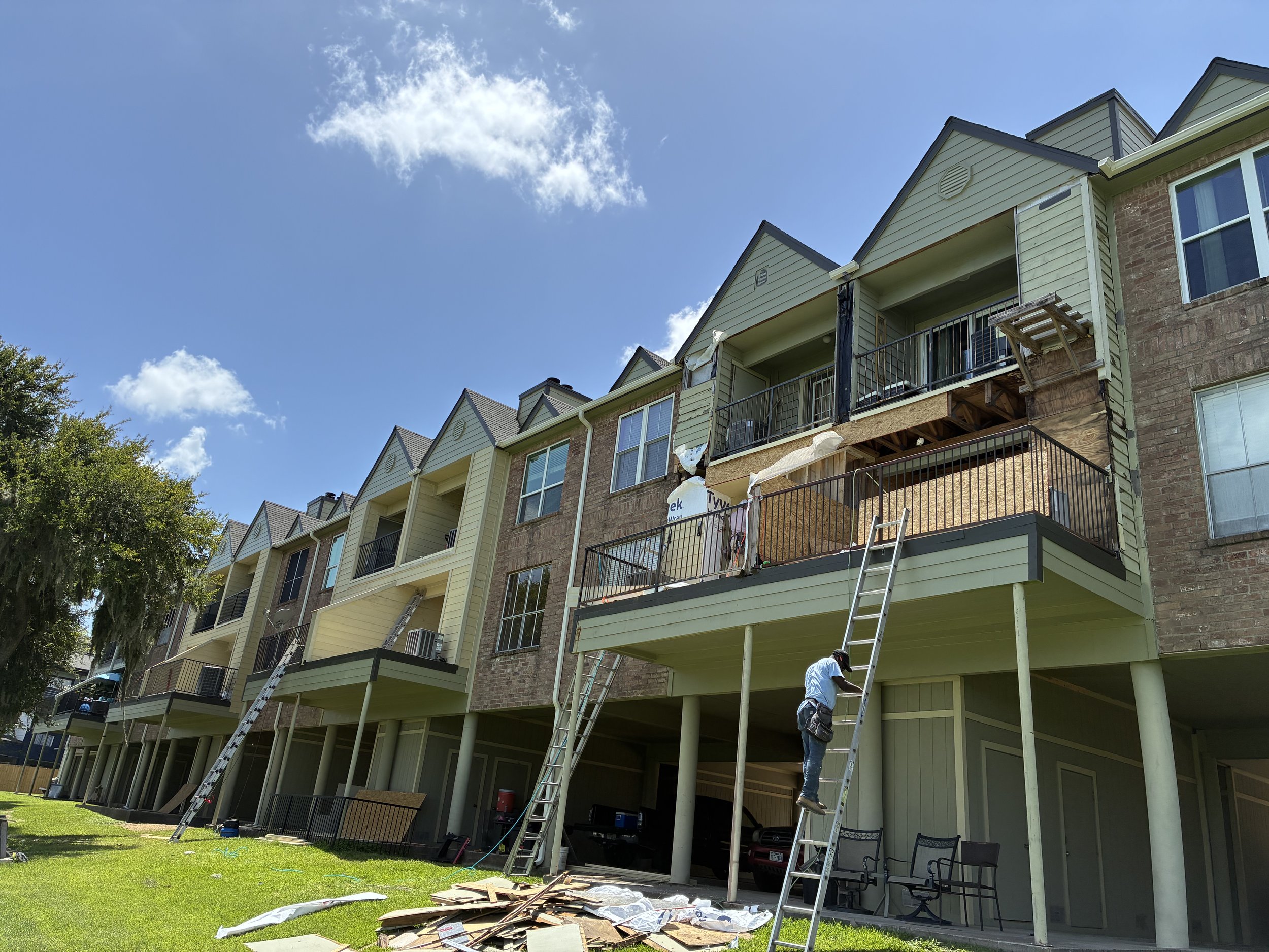 Construction workers are working on the balconies of an apartment complex, installing or repairing the railings. Ladders are leaning against the building, and construction materials are scattered on the ground beneath a sunny, clear sky.