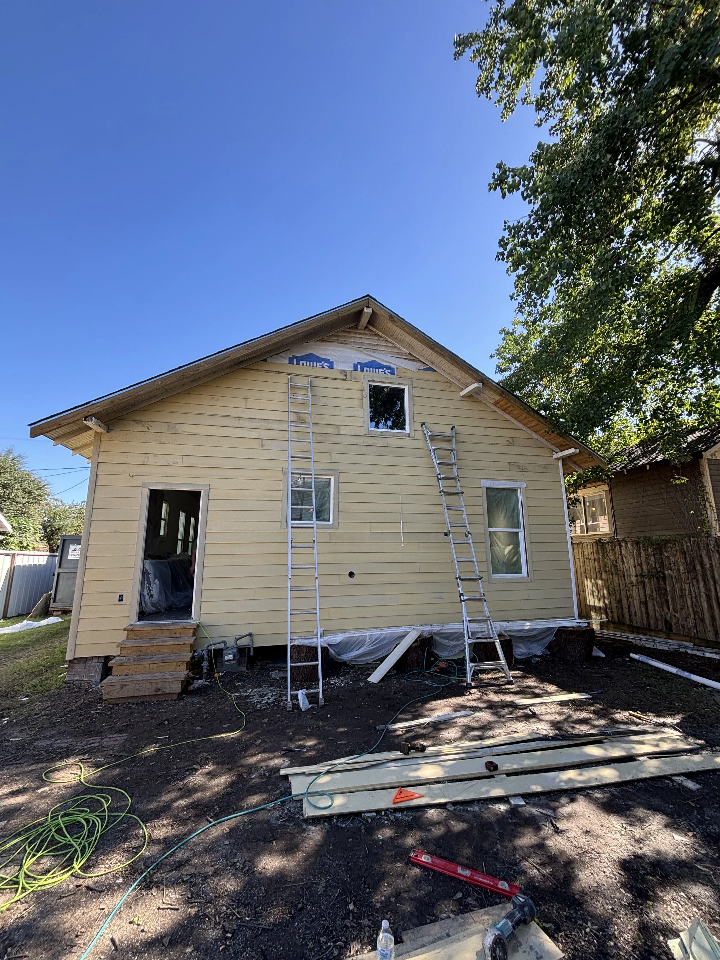 Under construction house with completed yellow siding, two aluminum ladders leaning against it, and some construction tools and materials on the ground, including a level, planks, an extension cord, and a power drill. The house has a new window and a