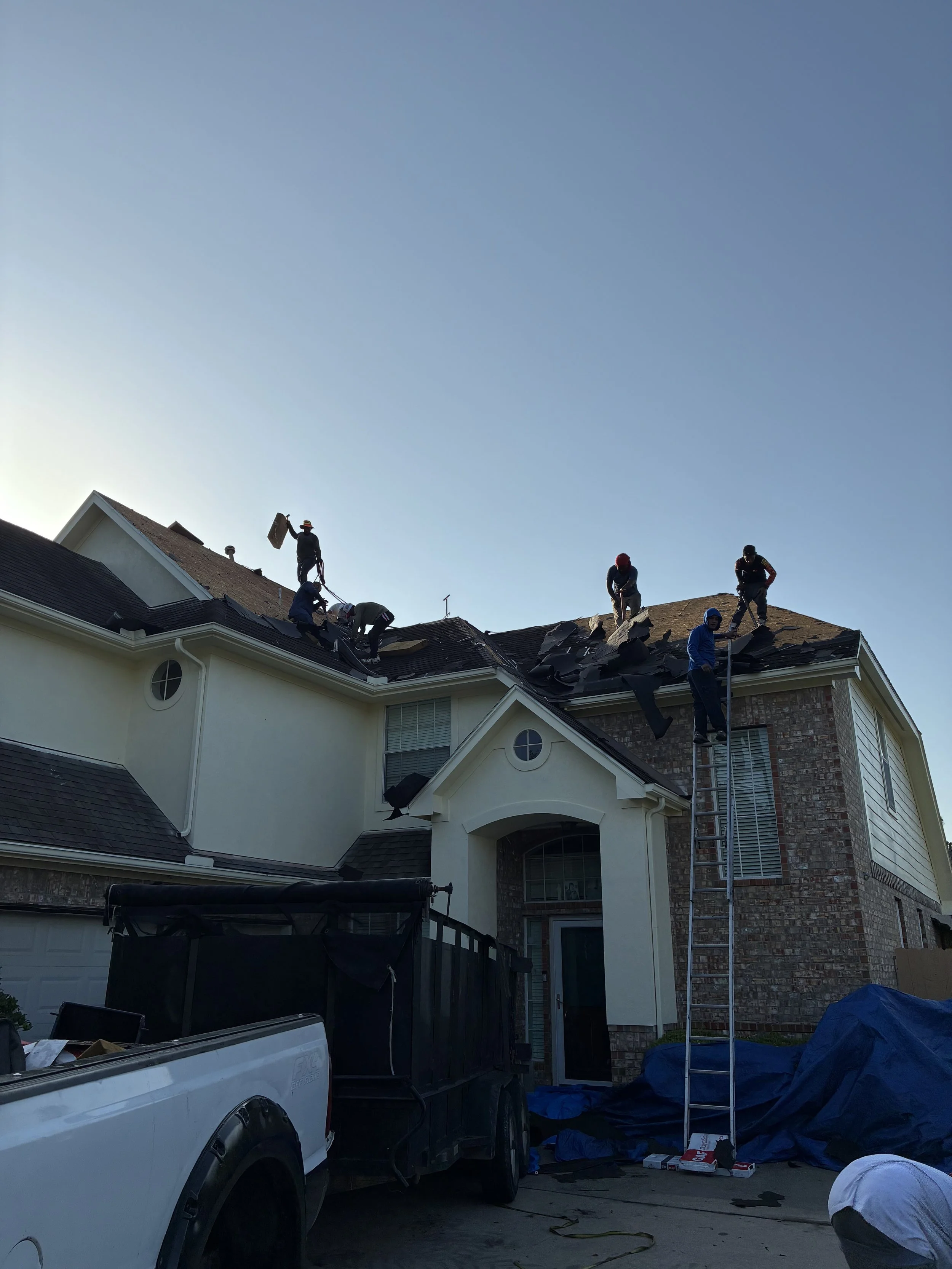 Workers on the roof of a two-story house, repairing or replacing shingles, with a ladder leaning against the house and a truck parked in the driveway.