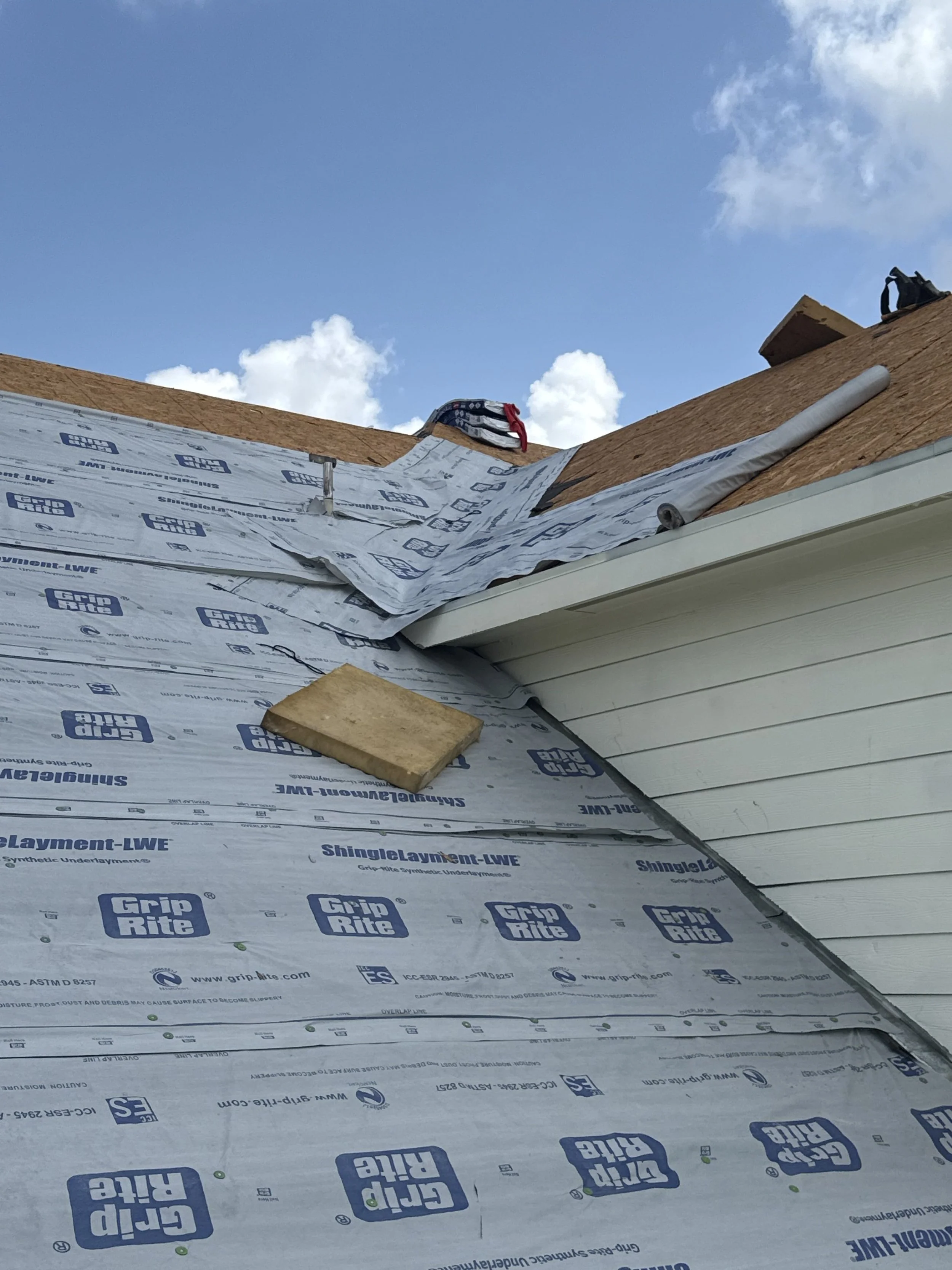 Construction site roof with underlayment material, wooden planks, and shingles in progress, under a partly cloudy sky.
