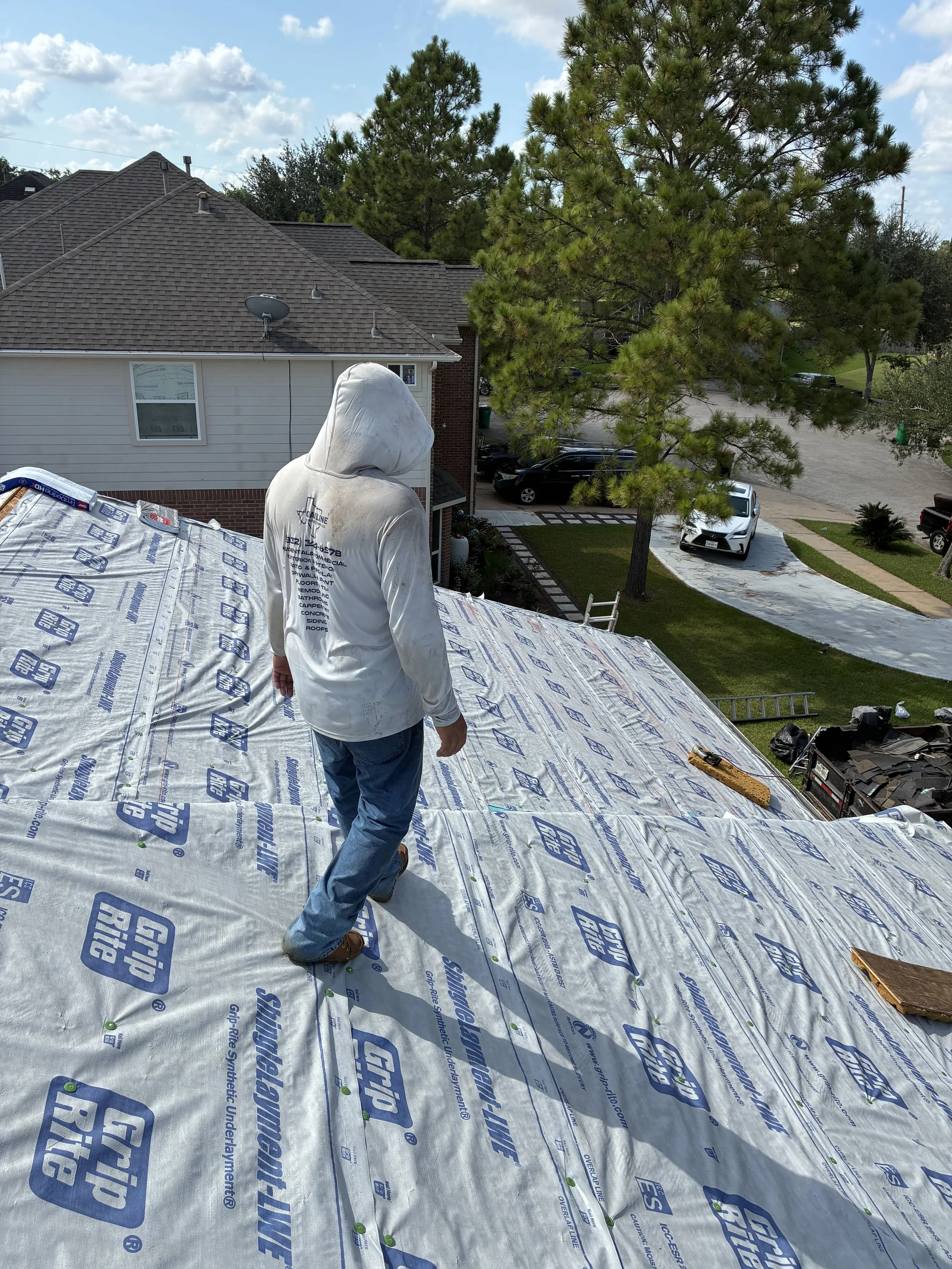 Person in a white hoodie walking on a roof under construction, with residential houses, trees, and parked cars visible in the background.