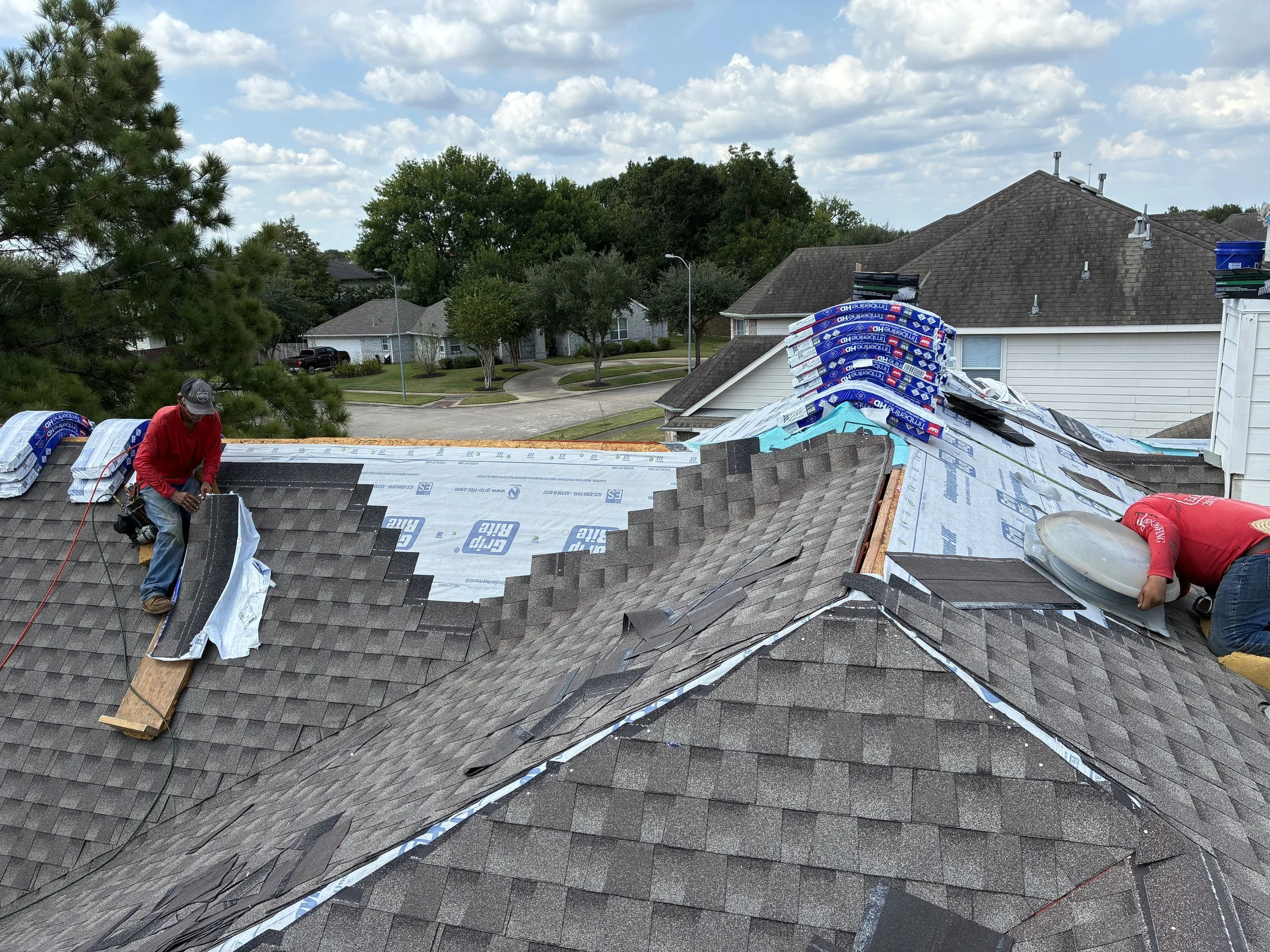 Two workers installing new shingles on a house roof during the daytime. One worker is on the left with a red shirt, and the other is on the right crouched down with a red shirt, handling a piece of roofing material. The roof has partially new shingle