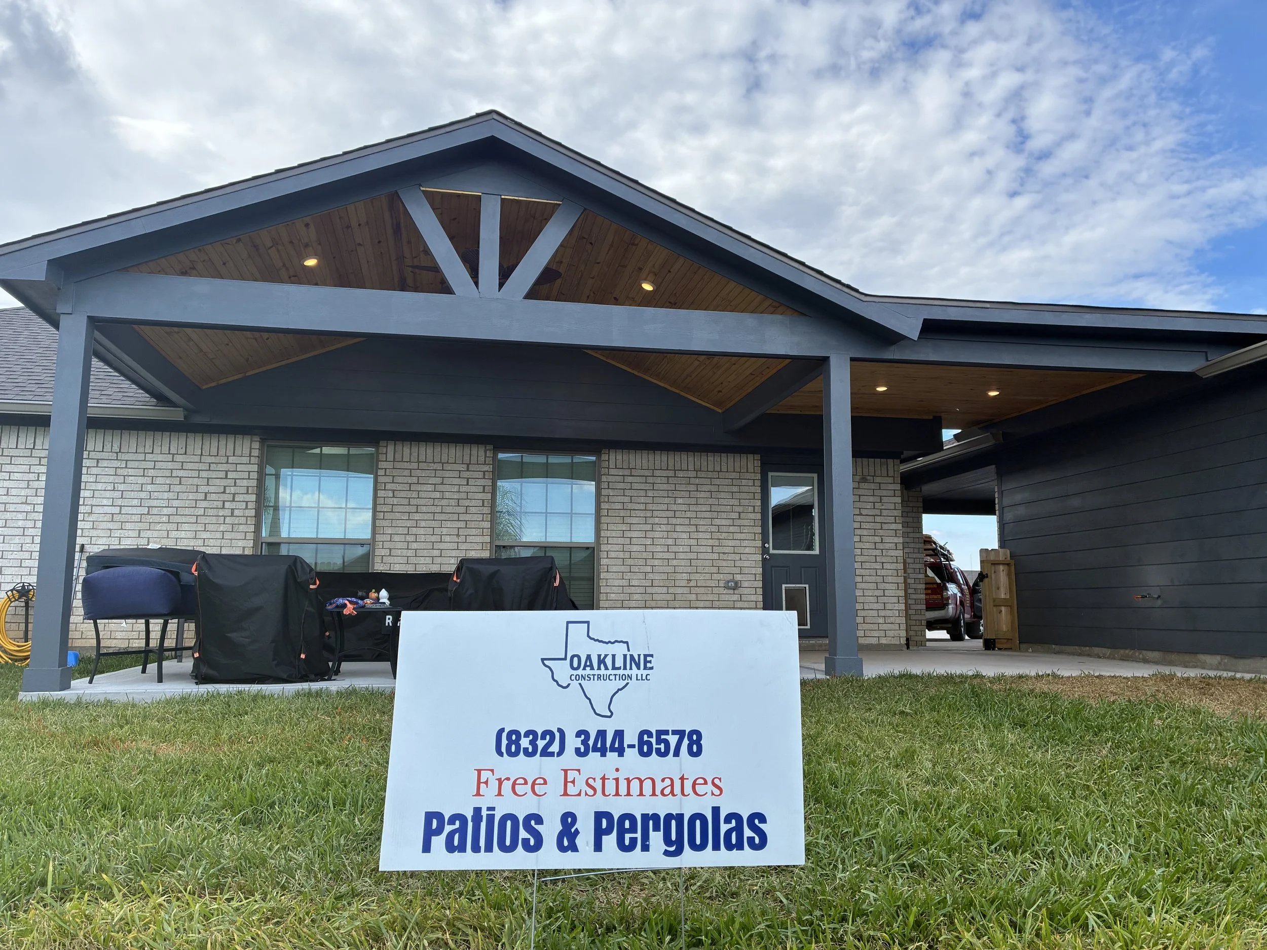 A house with a covered patio with a pergola, brick and dark wood siding, two windows, and a door. There is a sign on the grass advertising patios and pergolas by Oakline Construction LLC with a phone number for free estimates.