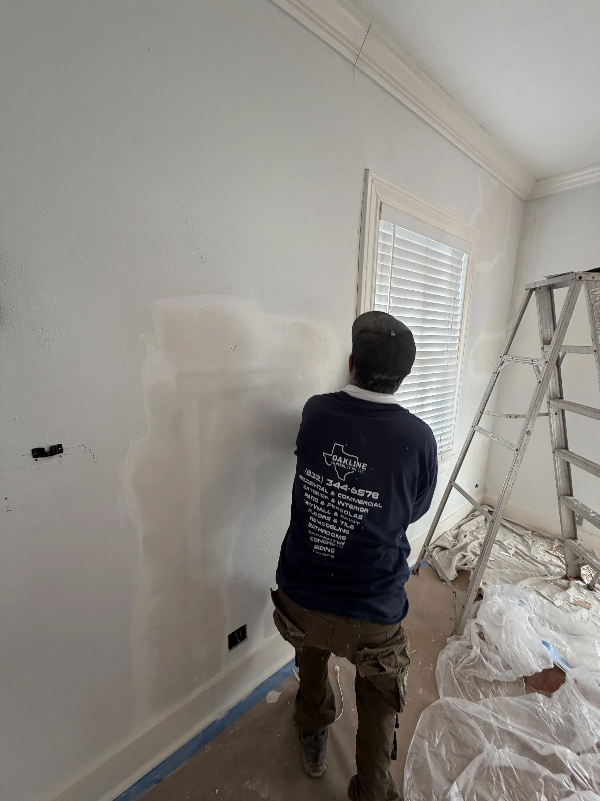 A person painting a wall in a room, standing with their back to the viewer, next to a ladder and window with blinds, with painting supplies and protective sheets on the floor.