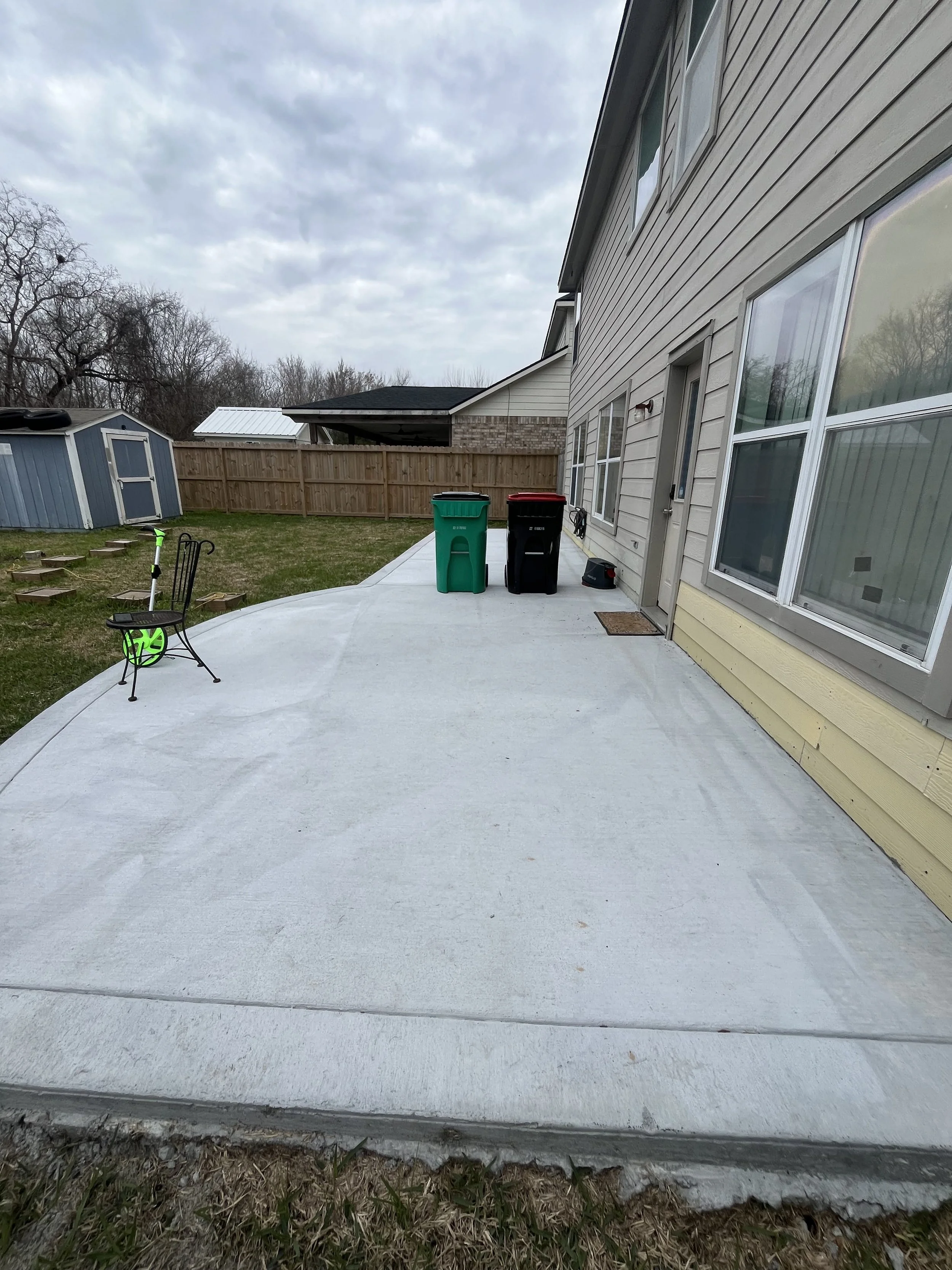 A backyard patio with a concrete surface, a green trash bin, a black trash bin, a chair, a small scooter, and a hose reel, with a house, lawn, and fenced yard in the background under cloudy skies.