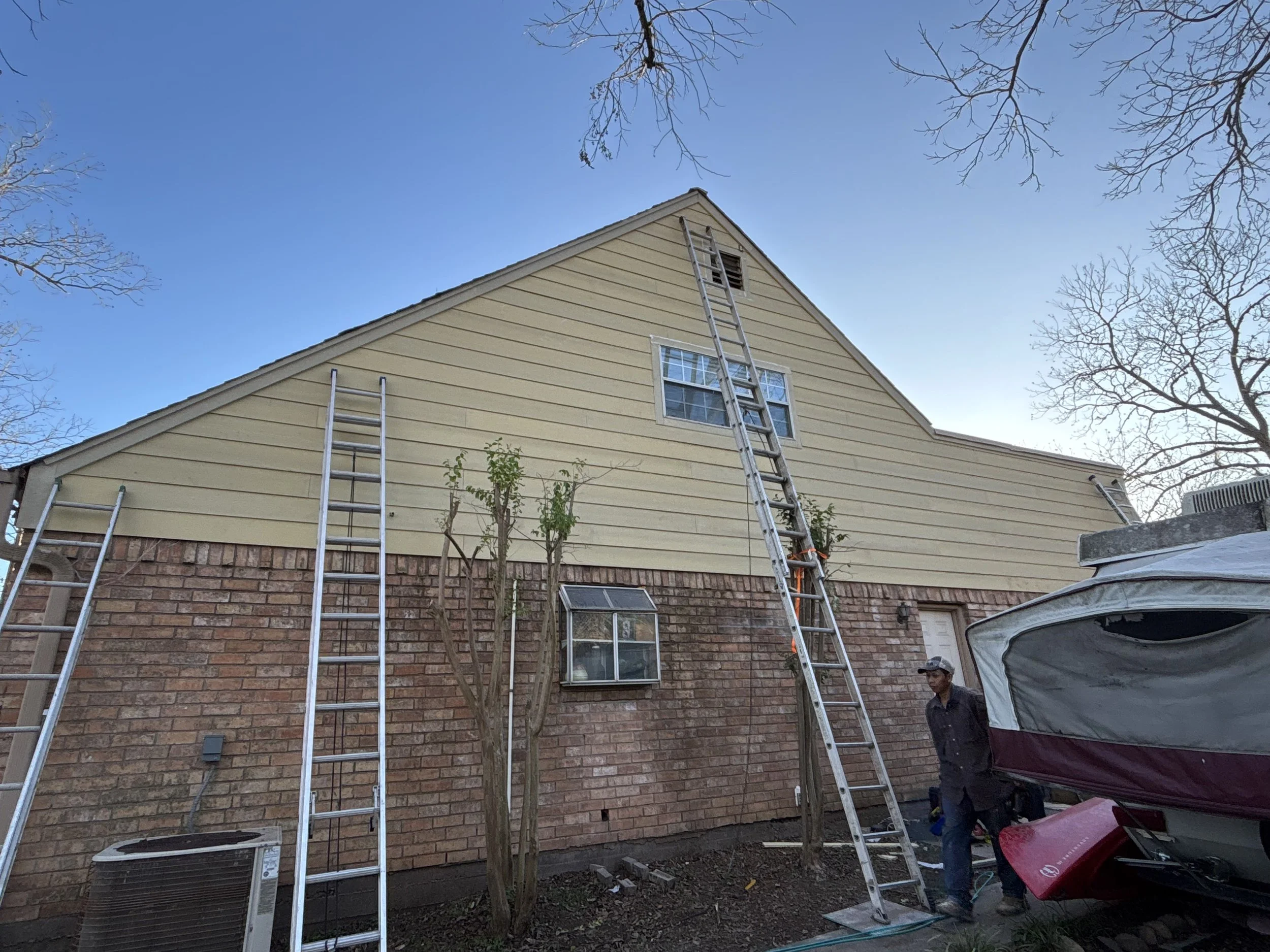 Side view of a house under renovation with three ladders leaning against the exterior wall, one reaching the roof. A person wearing a dark shirt and jeans stands near a boat trailer in the yard. Tall trees with bare branches are seen, and the sky is 