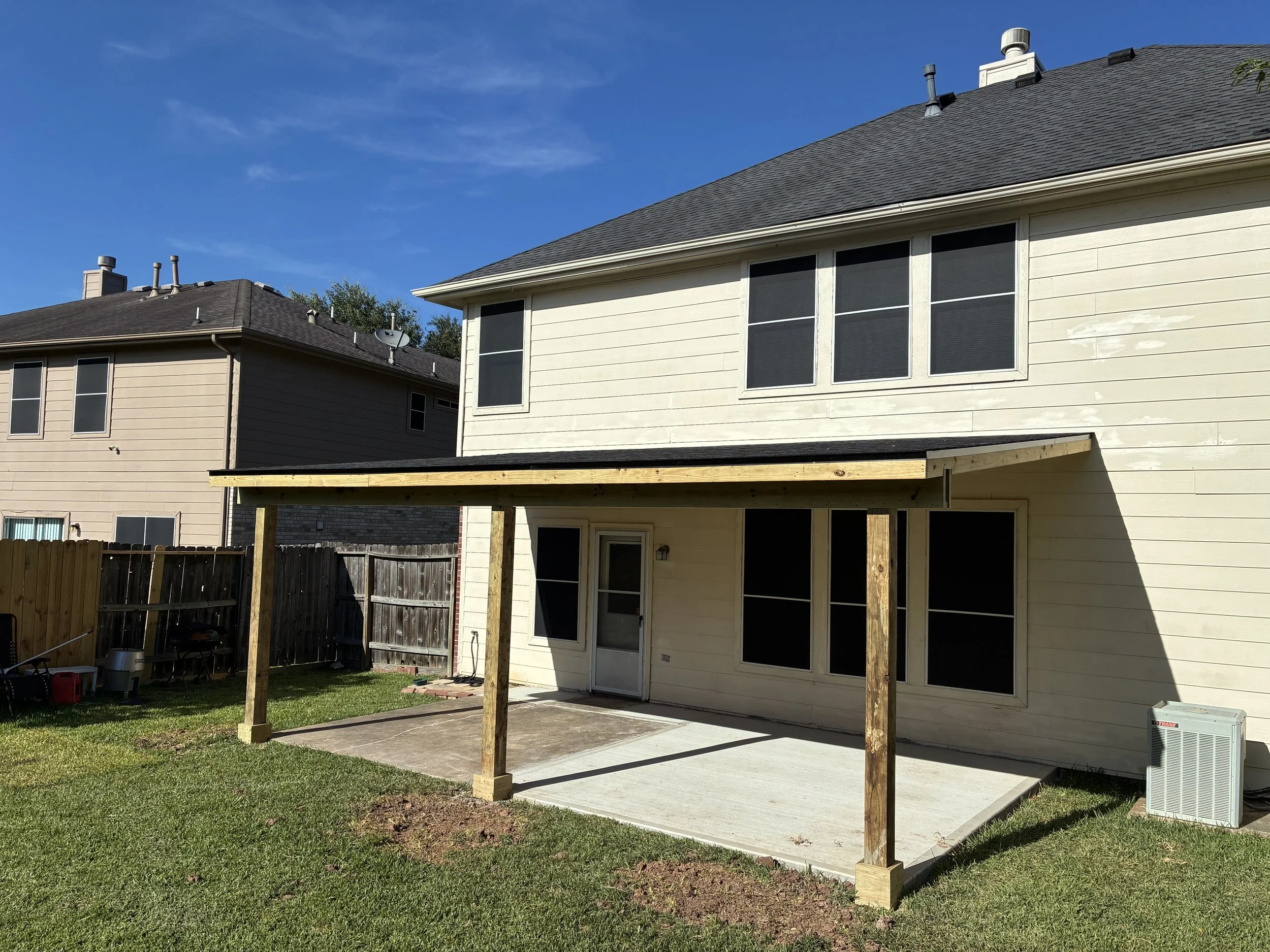 Backyard with a newly built wooden patio cover extending from the house, with a concrete slab floor, surrounded by green grass and a wooden fence, under a clear blue sky.