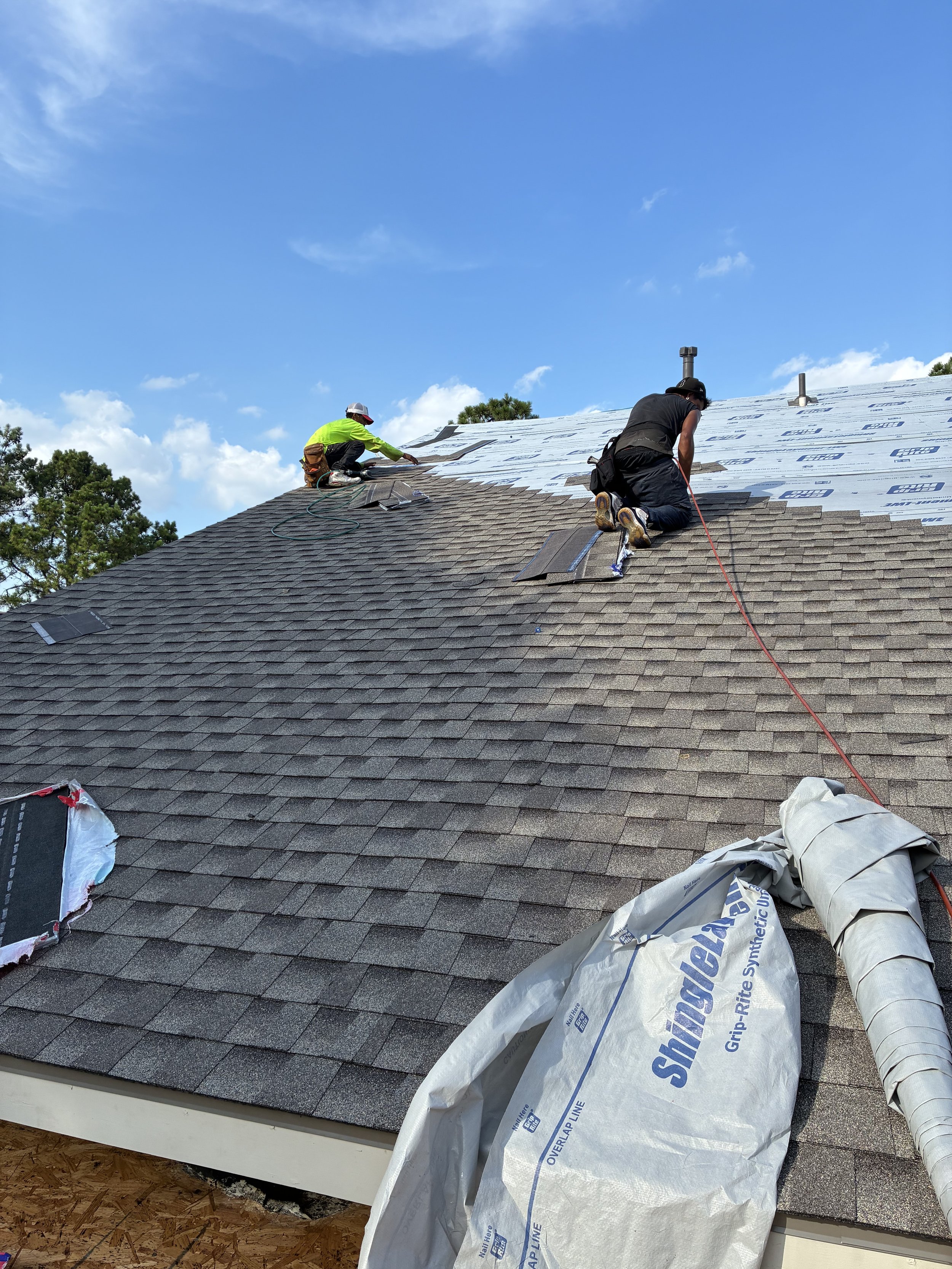 Two workers on a sloped roof installing shingles, with one in a yellow shirt and the other in black, under a blue sky with some clouds.