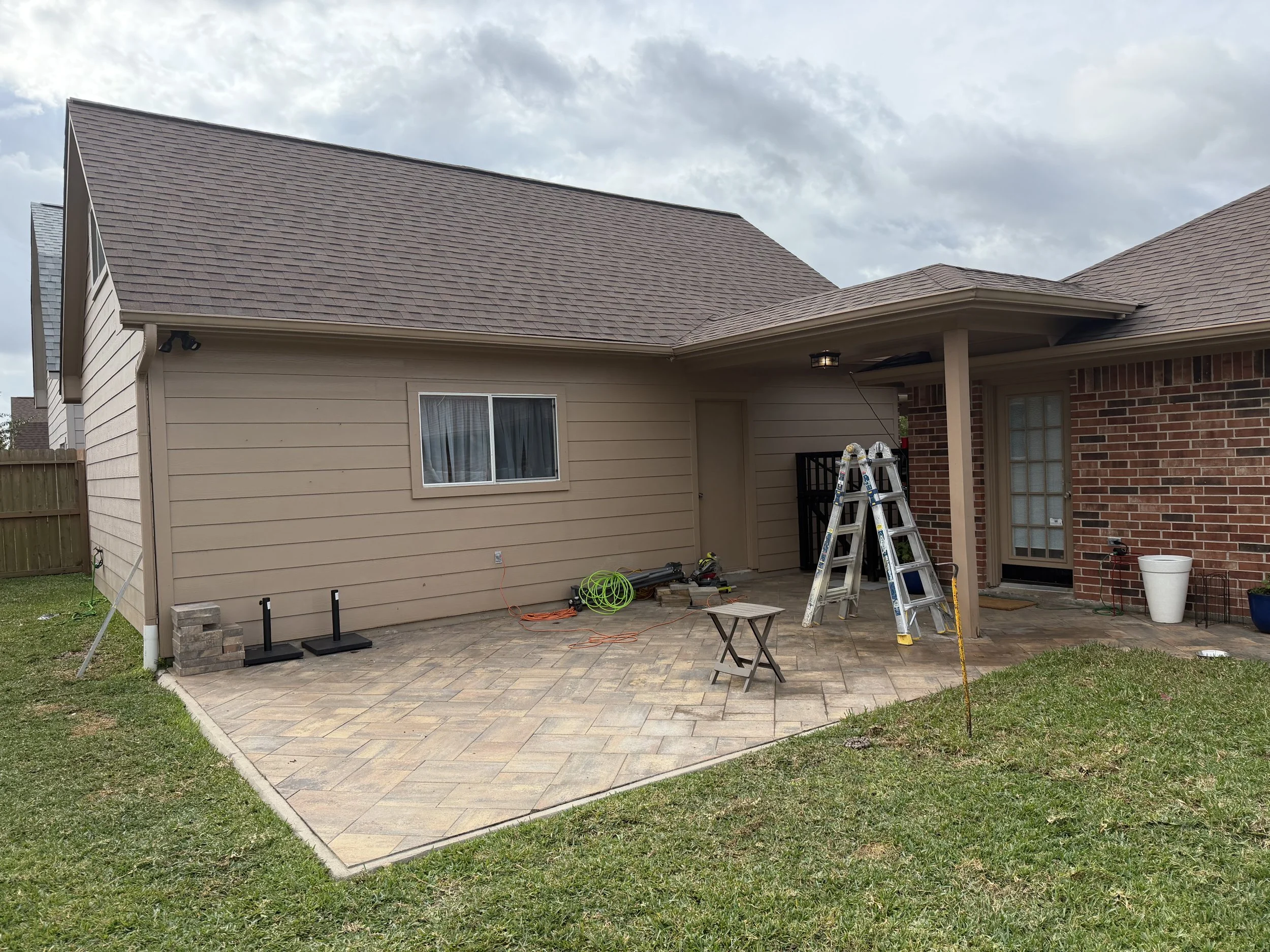 Back patio of a house with a brick and beige siding exterior, displaying construction tools including a ladder, small table, and various equipment, with a cloudy sky overhead.