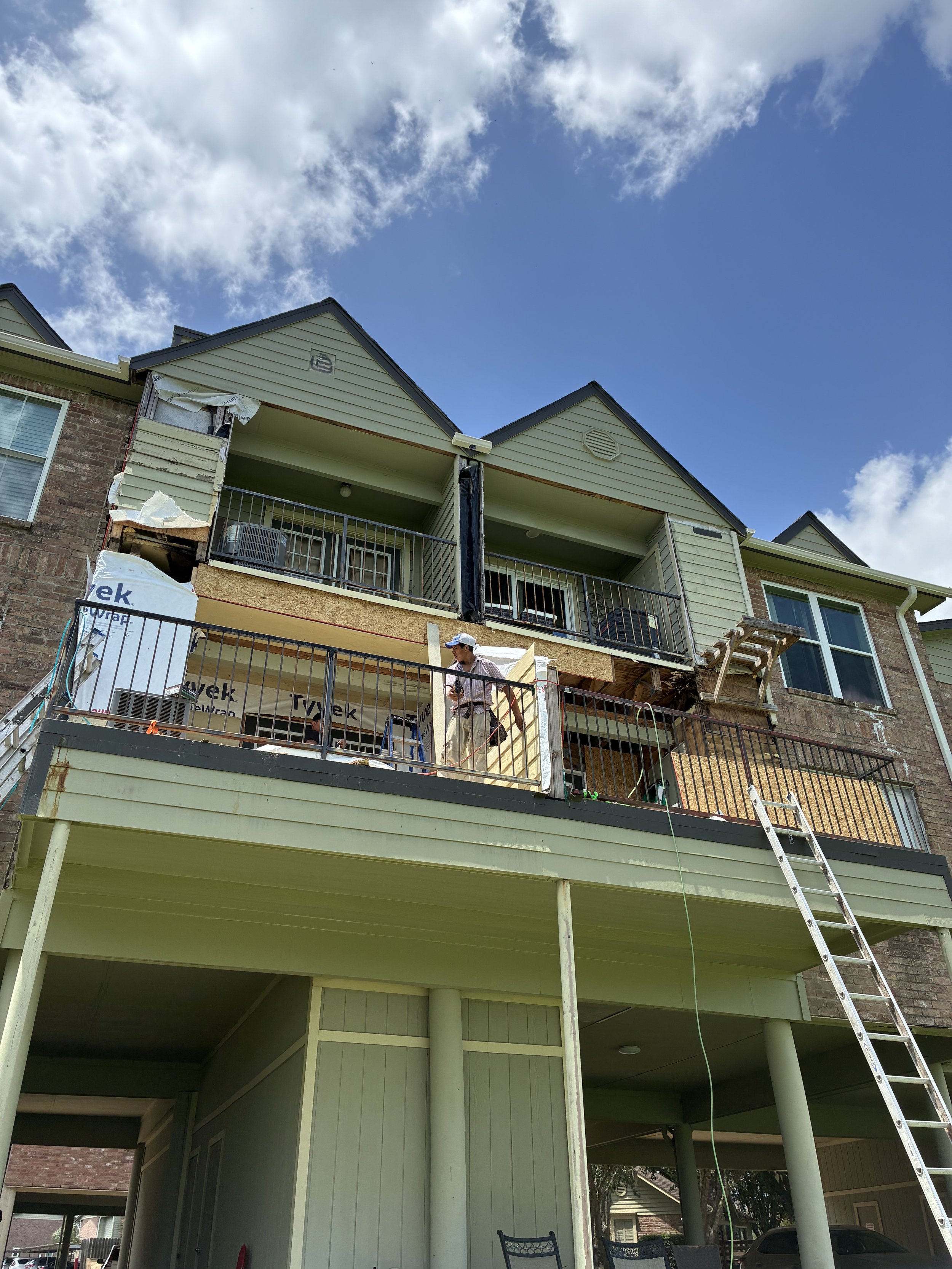 Construction workers renovating the exterior of a multi-story residential building. The upper balcony areas are being upgraded with new railing and siding. A ladder is positioned on the right side of the building, and construction materials and tools