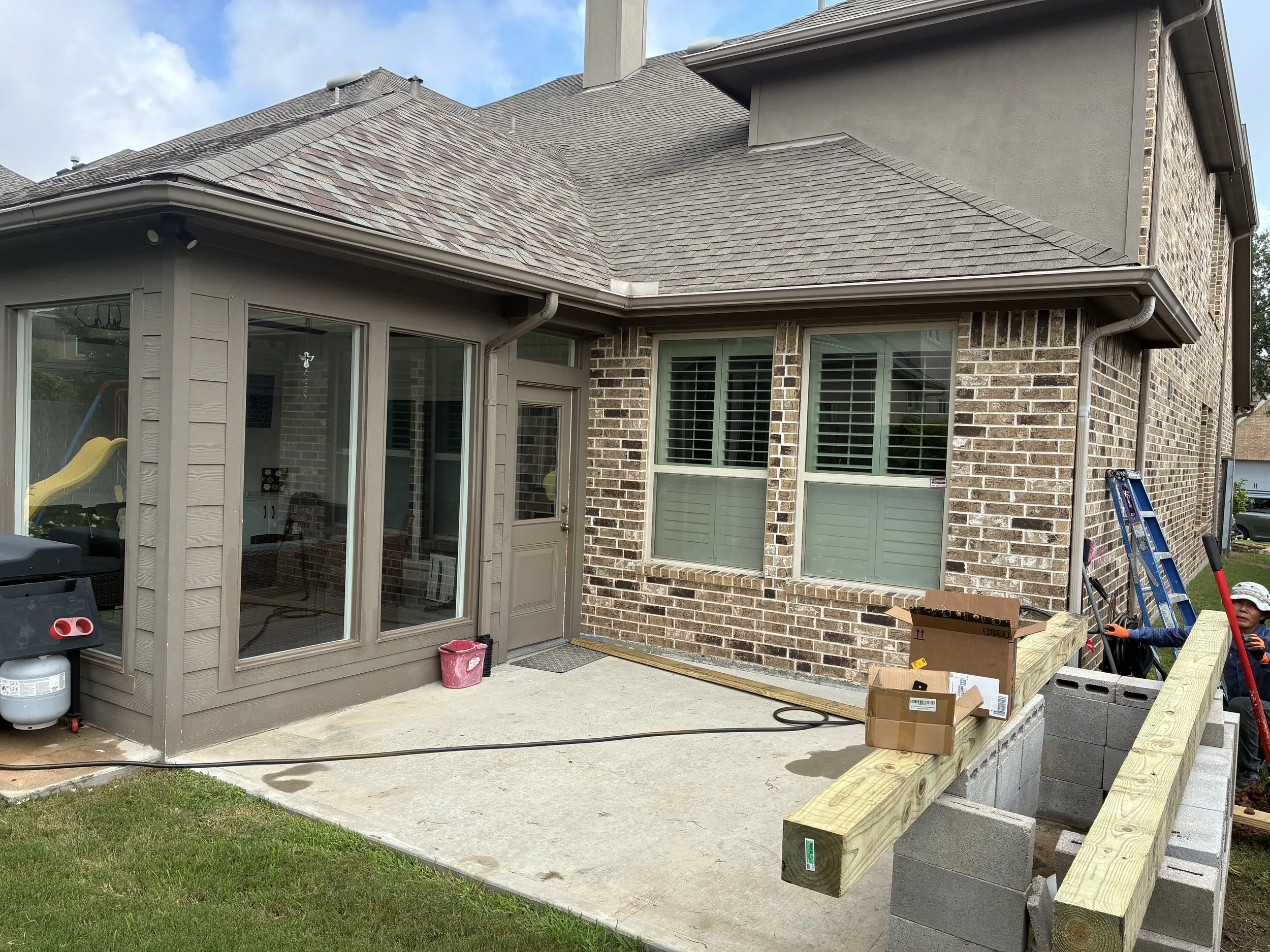 Construction site of a backyard patio extension with cinder blocks and wooden beams, next to a brick house with large windows and sliding glass door, a man in safety gear working, ladder, open cardboard boxes, and a grass lawn.