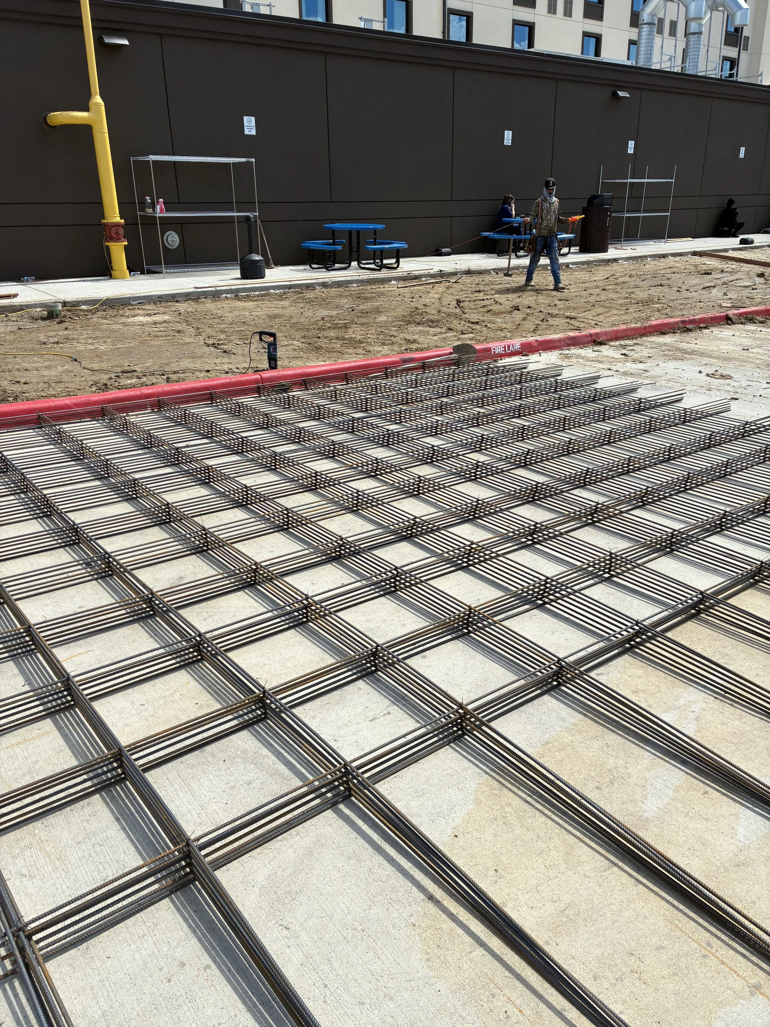 Construction site showing steel rebar grid on the ground with workers in the background and some outdoor furniture near a dark wall.