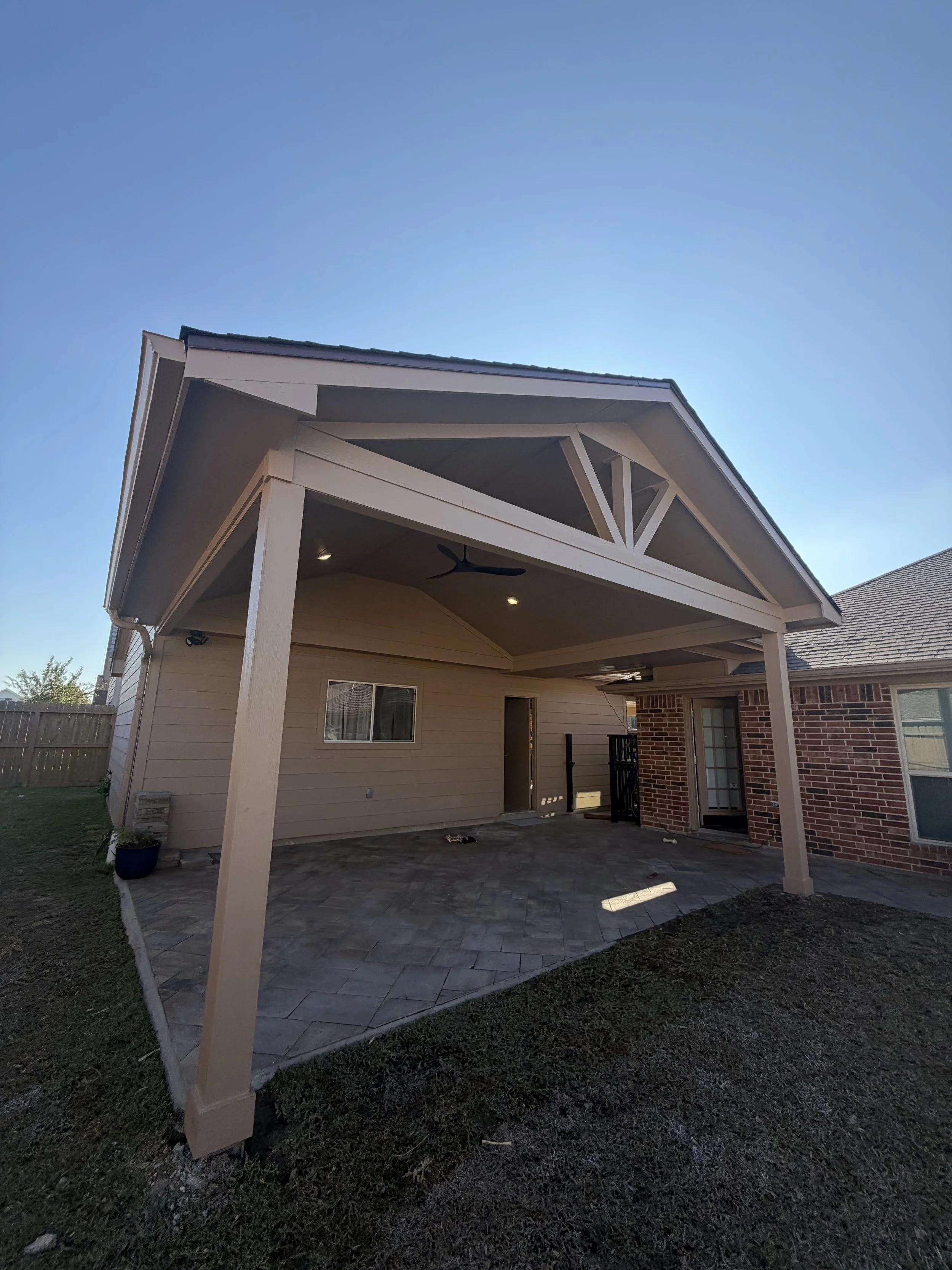 Back patio of a house with a beige covered porch, paver floor, hanging ceiling fan, and brick wall outdoor door.