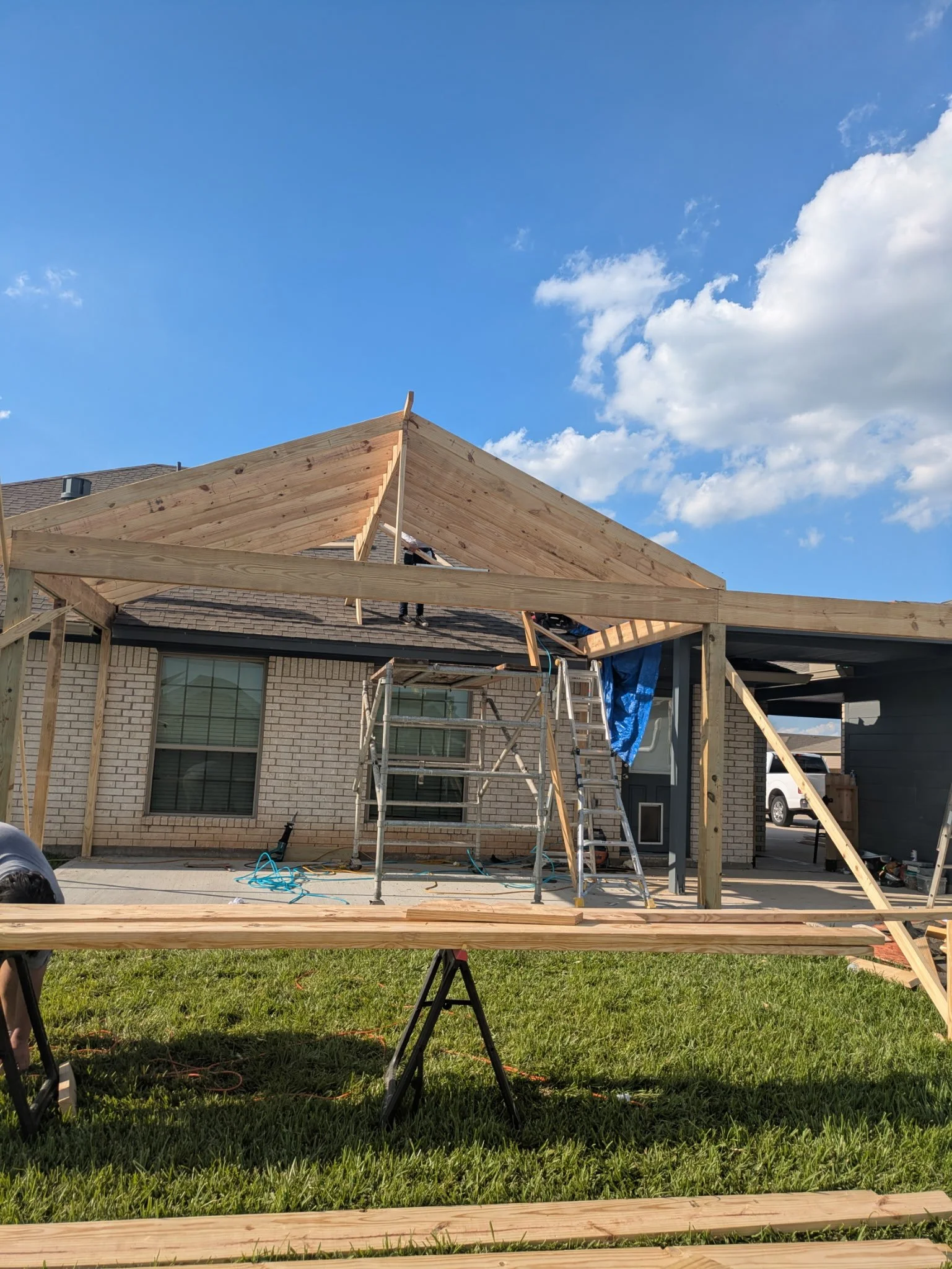 Construction workers building a roof extension on a brick house, with the framework and roof trusses partially installed, set against a blue sky with some clouds.