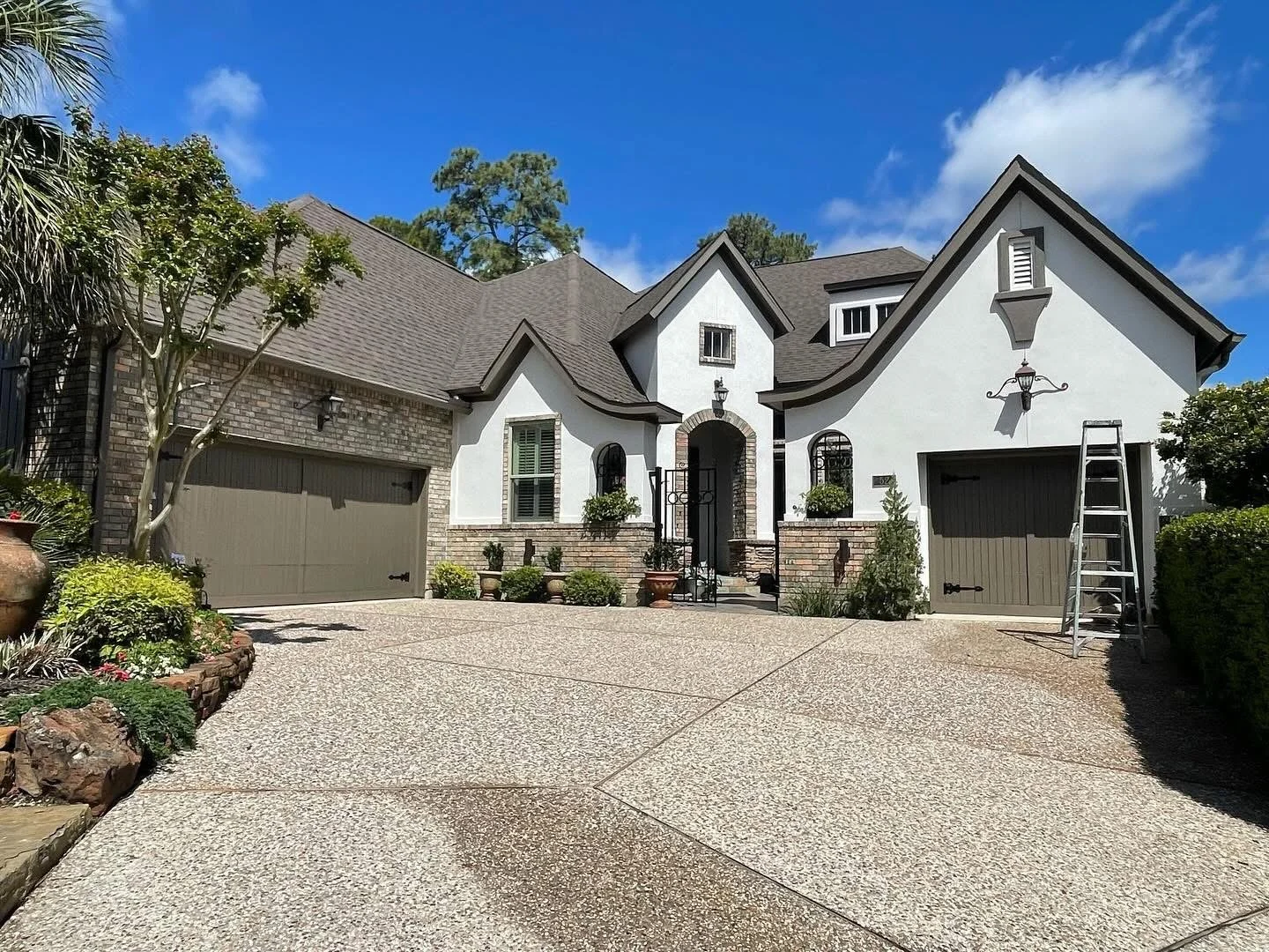 A large white house with a gray roof, two attached garages, and a small front garden with plants and potted flowers. The driveway is made of textured concrete and a ladder leans against the right garage. The house has decorative lanterns and small wi