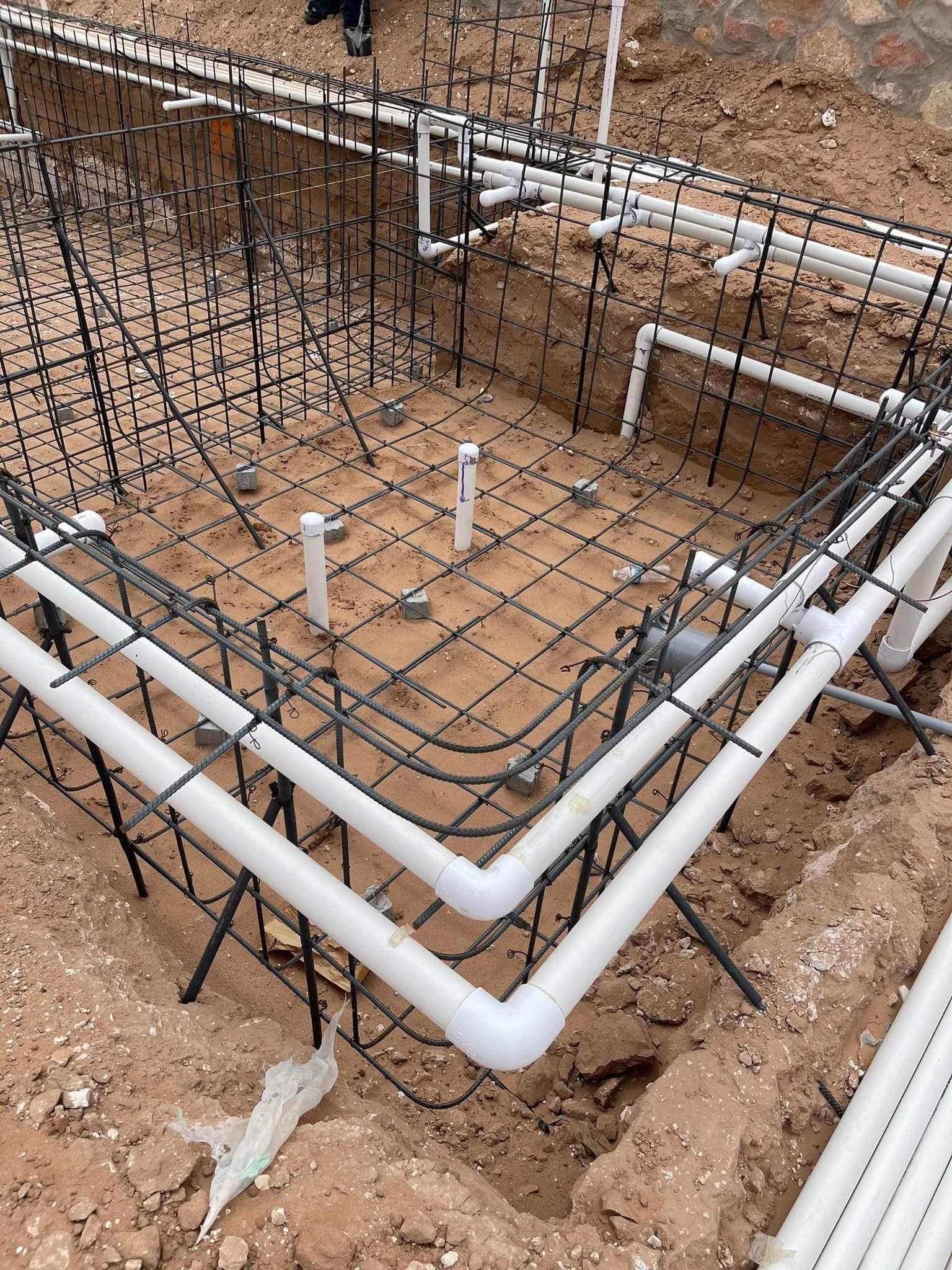 Construction site with metal rebar, black wires, and white PVC pipes installed in a rectangular foundation.