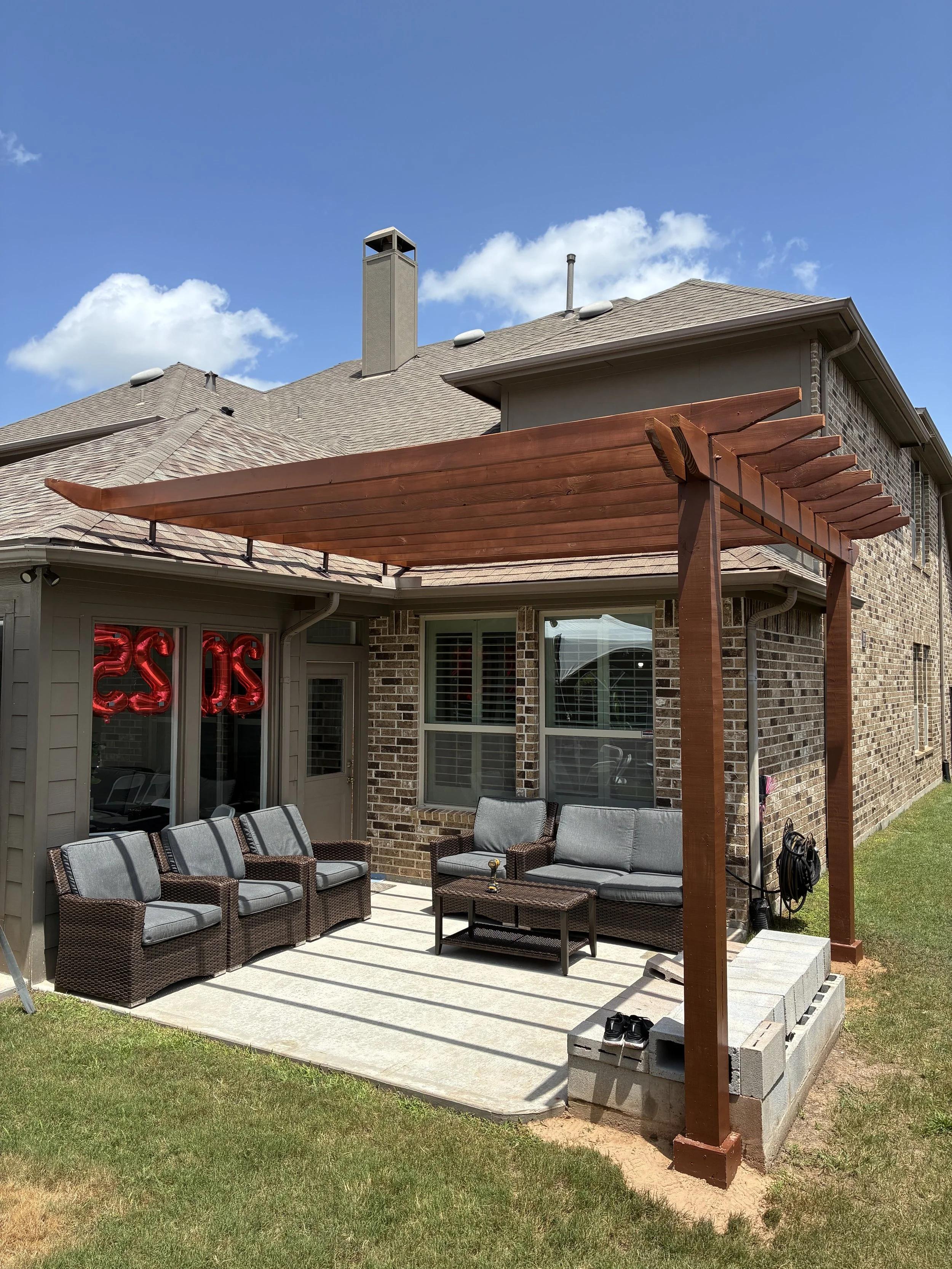 Backyard patio with outdoor furniture, including a sofa, chairs, a table, and a wooden pergola, next to a brick house with a window and a door.