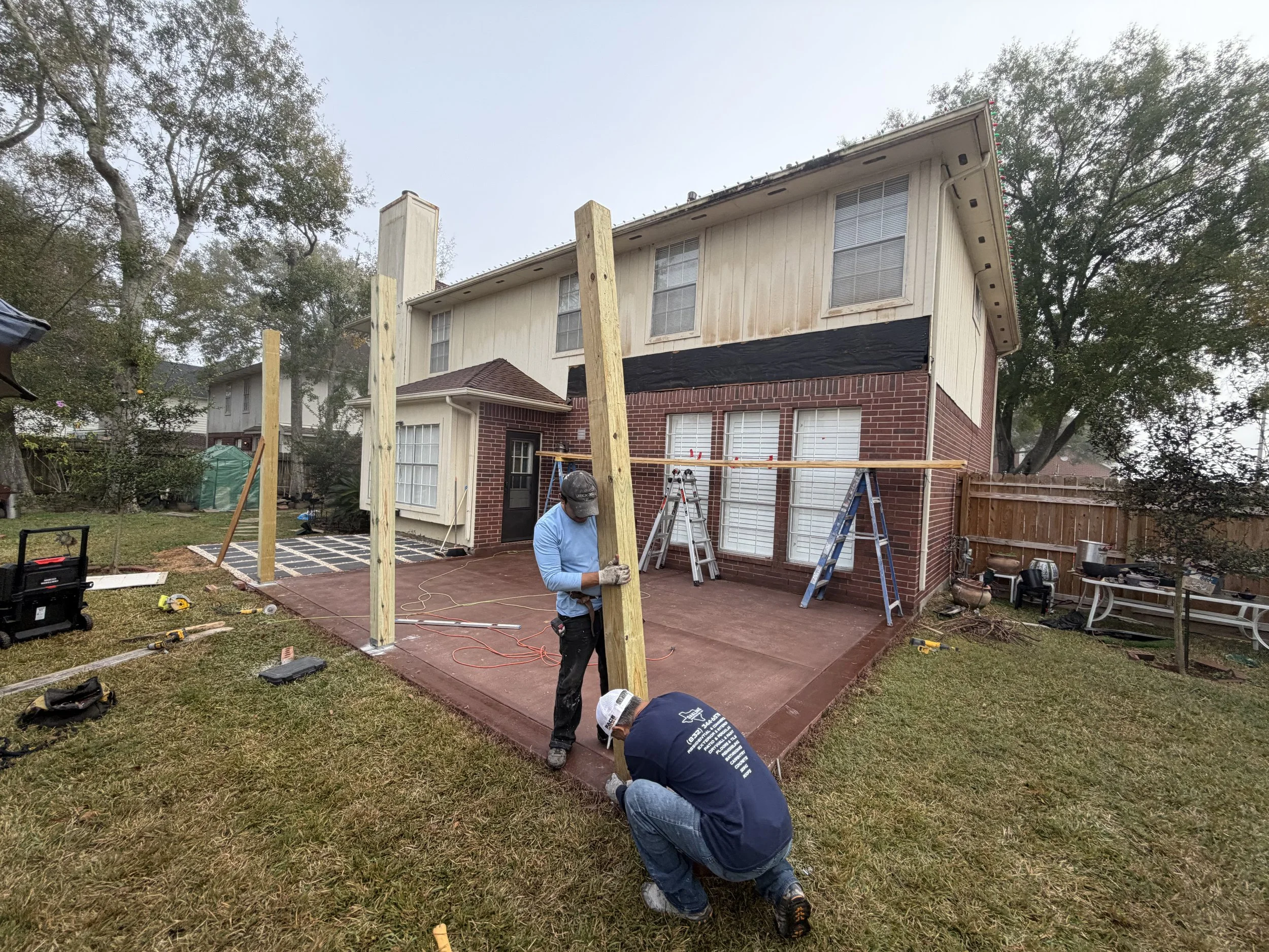 Two workers assembling wooden posts on a backyard patio, preparing for a fence installation. There are ladders, tools, and construction materials around.