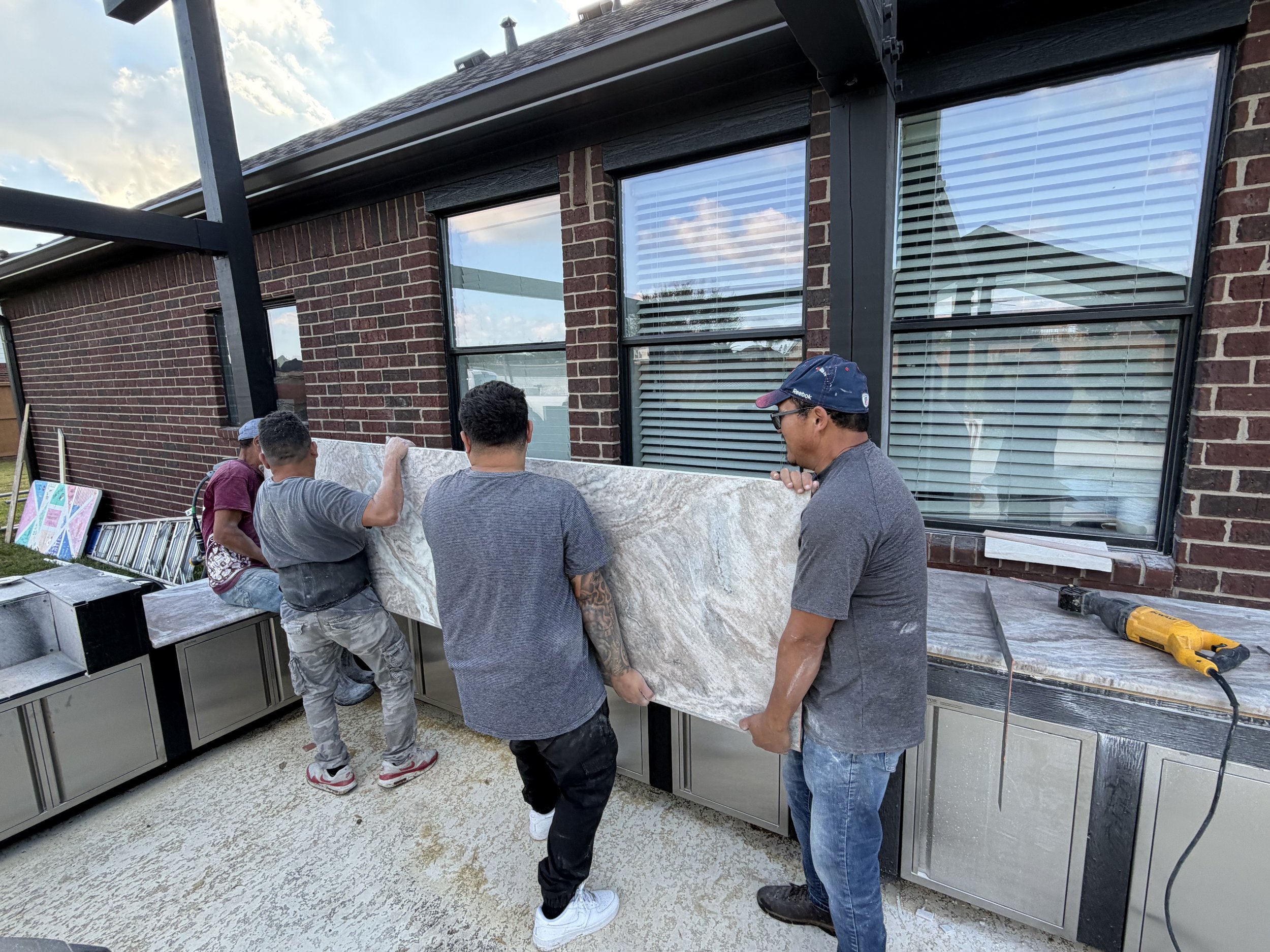 Group of four men installing large marble or granite slab on outdoor kitchen counter.