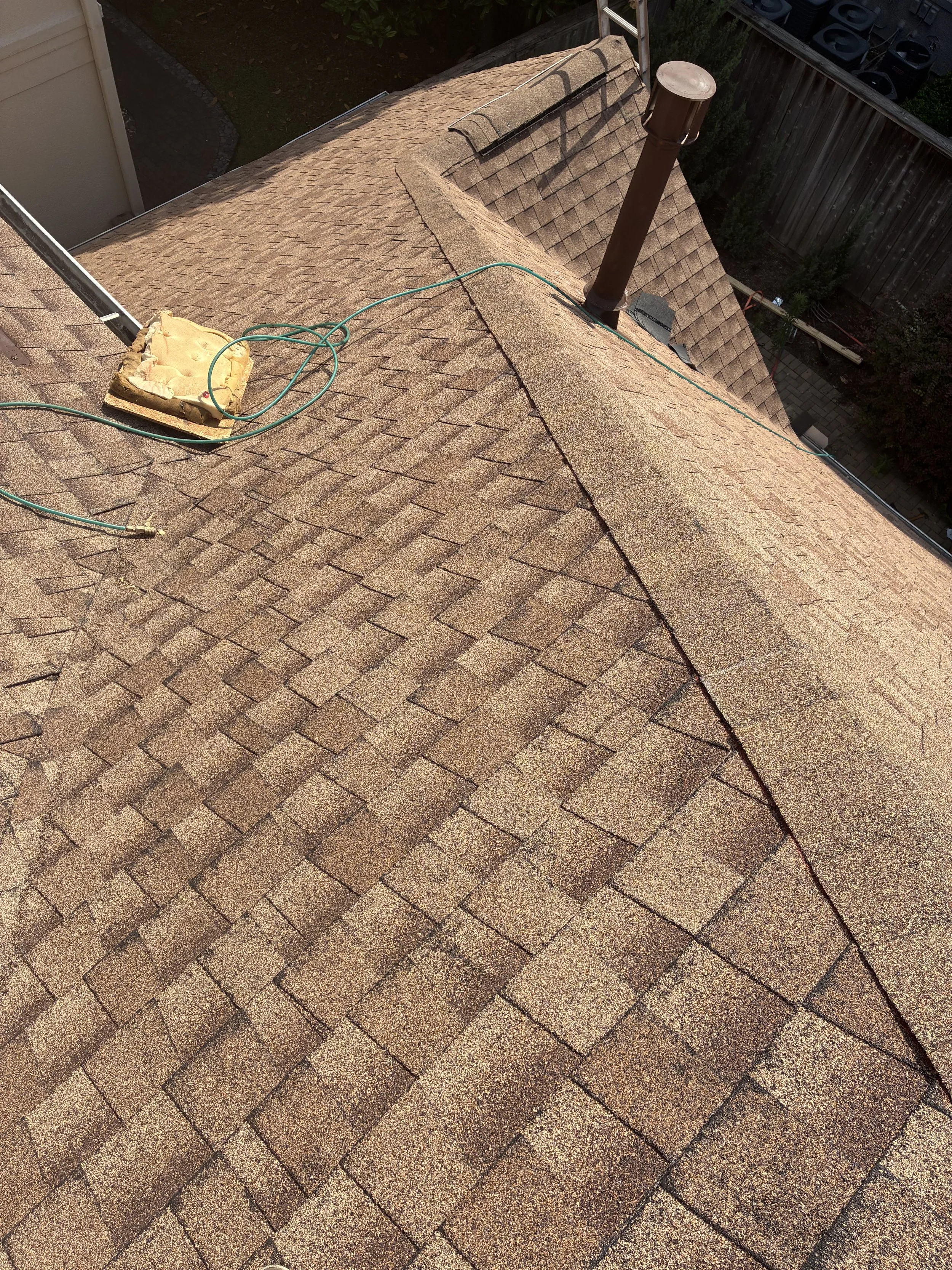 View of a residential shingle roof with a ridge cap, a vent pipe, and an electrical outlet on the roof, with neighboring houses and fences visible.