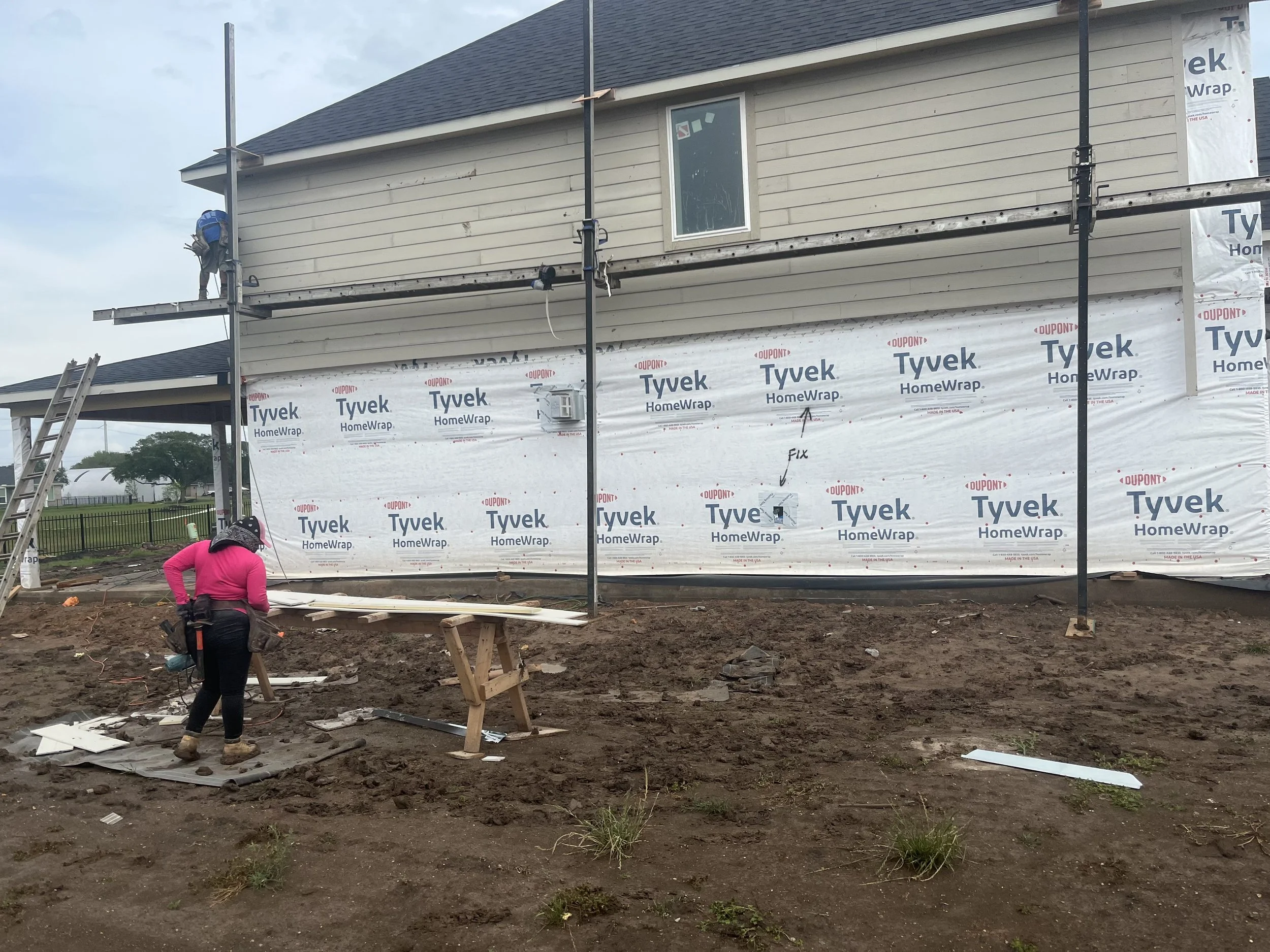 Construction worker in pink shirt and black pants working on the exterior of a house in the process of being built. The house is covered with Tyvek HomeWrap and has scaffolding and a ladder nearby.