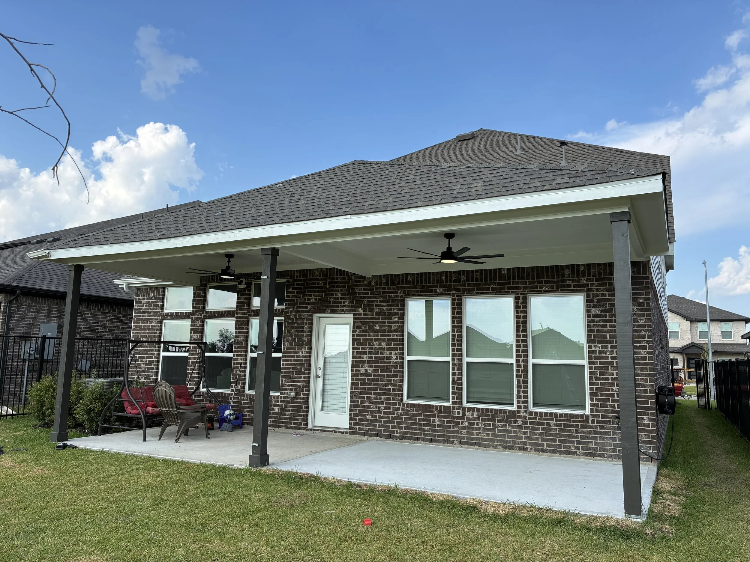 Backyard patio with a concrete floor, covered by a roof with ceiling fans. Features outdoor furniture including a swing and a plastic chair, surrounded by a lawn and enclosed by a black fence. A brick house with four windows and a white door is visib