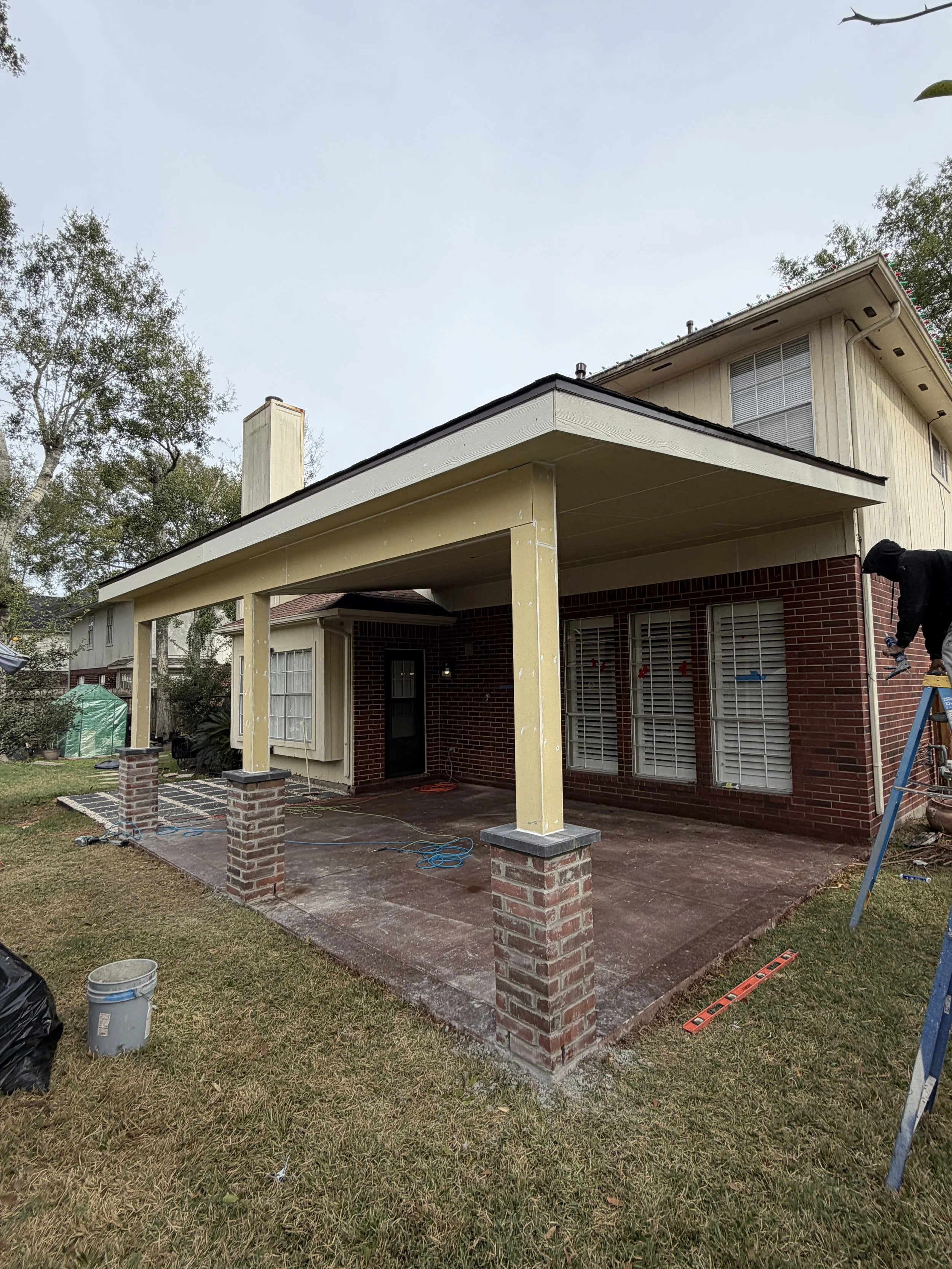 Construction workers working on a house porch, with columns being painted or repaired, and tools scattered around.