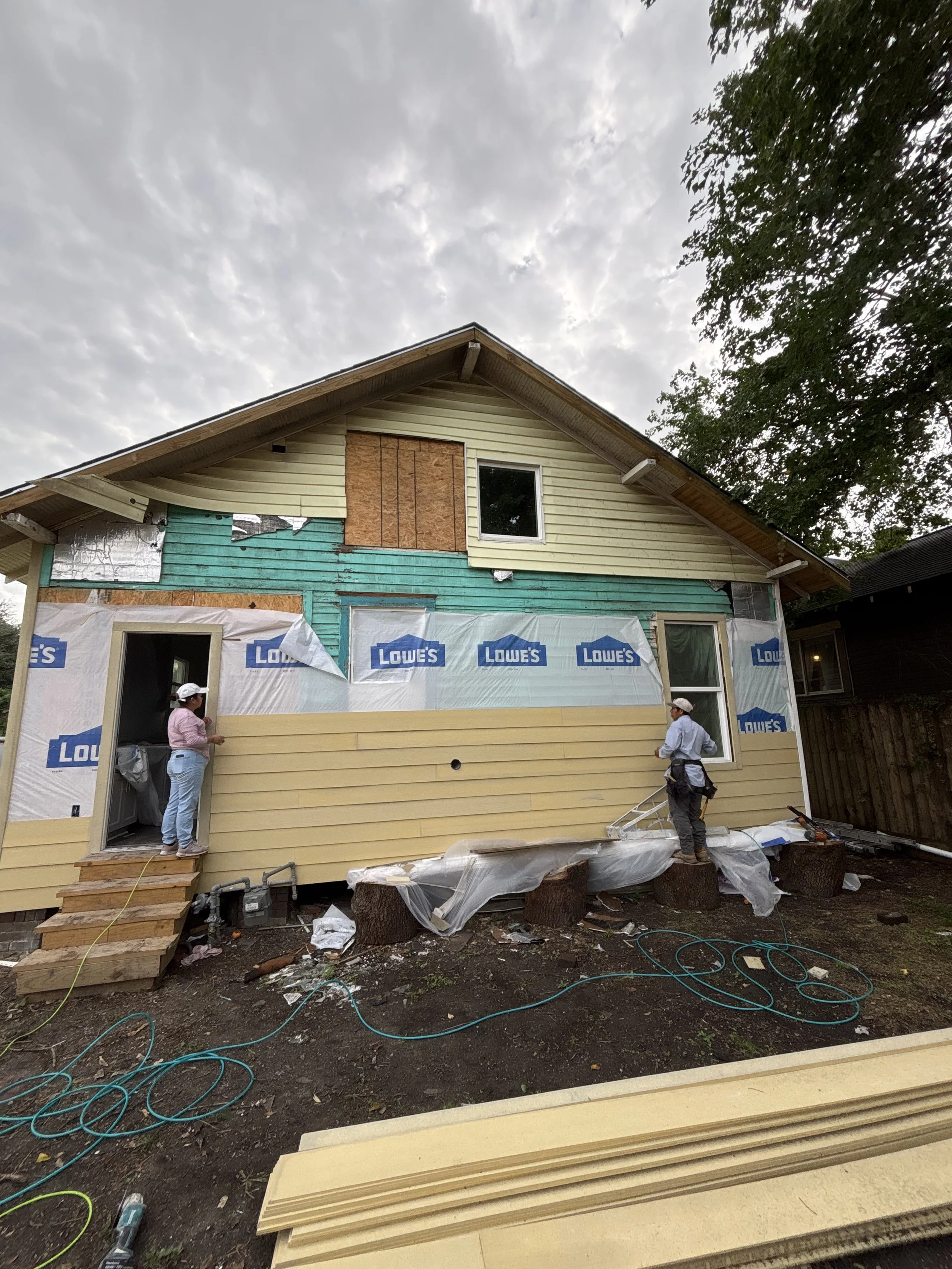 House undergoing renovation with workers installing siding, Lowe's insulation wrap visible, and construction materials scattered around.