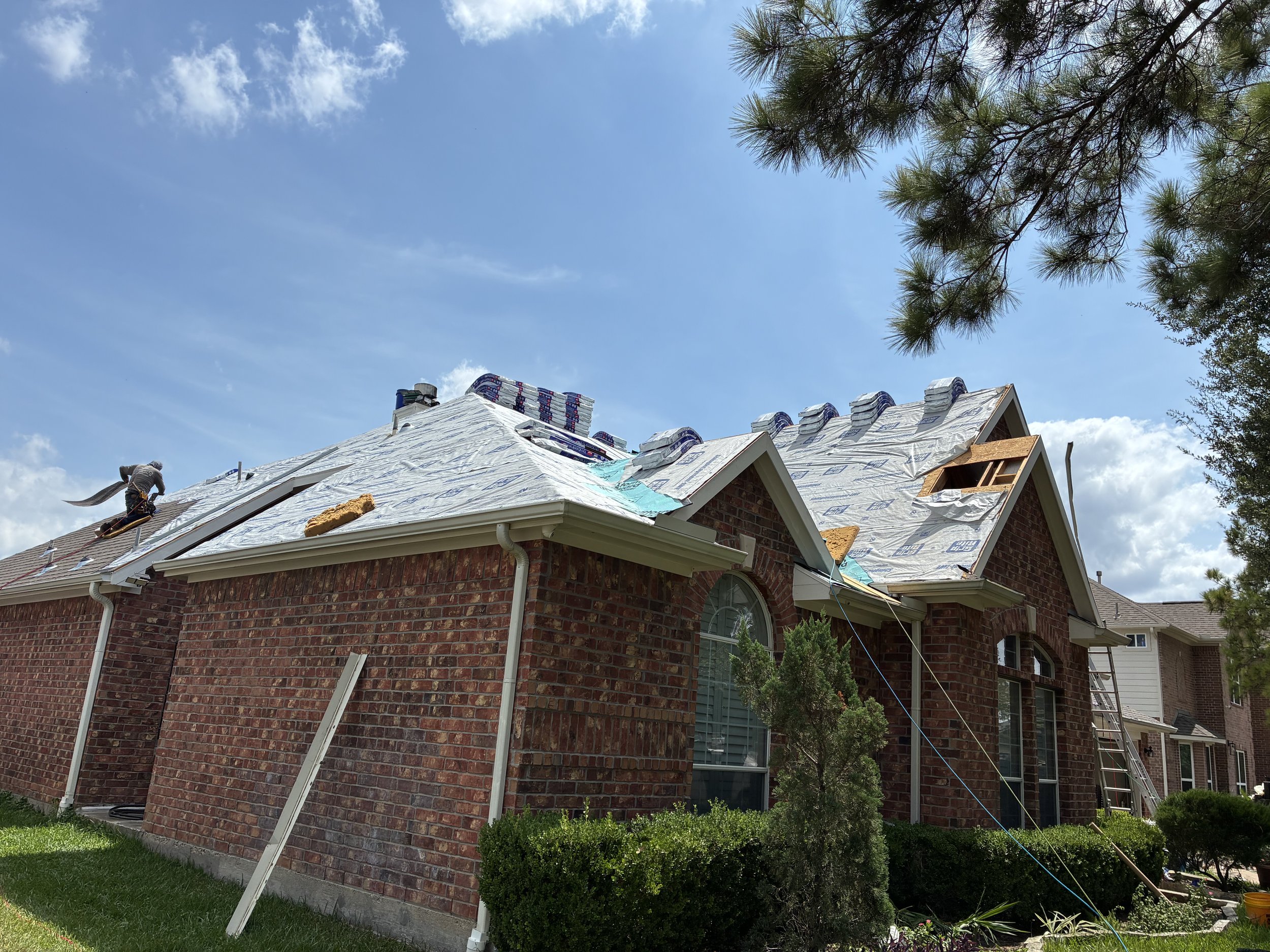 A brick house under construction or renovation, with workers on the roof installing new roofing materials, blue sky, and green trees surrounding the house.