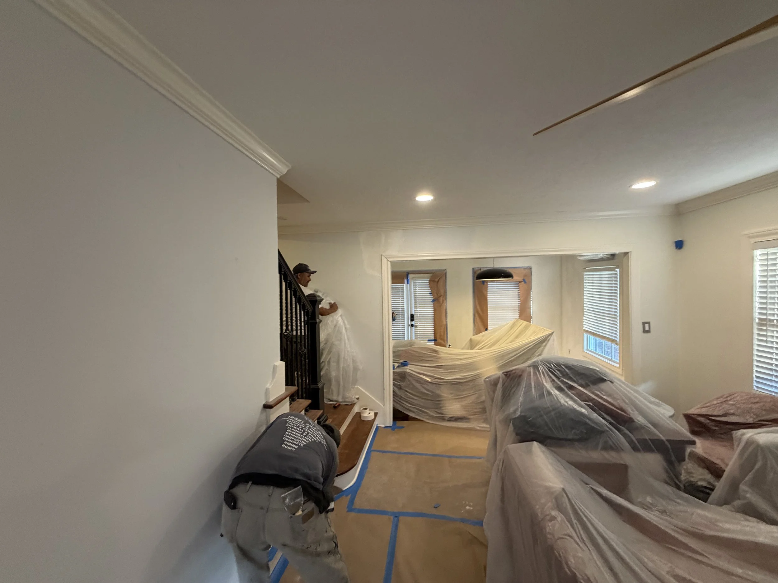 Living room undergoing renovation, with furniture covered in plastic, a person standing on the stairs, window blinds, and taped protective coverings on the walls.