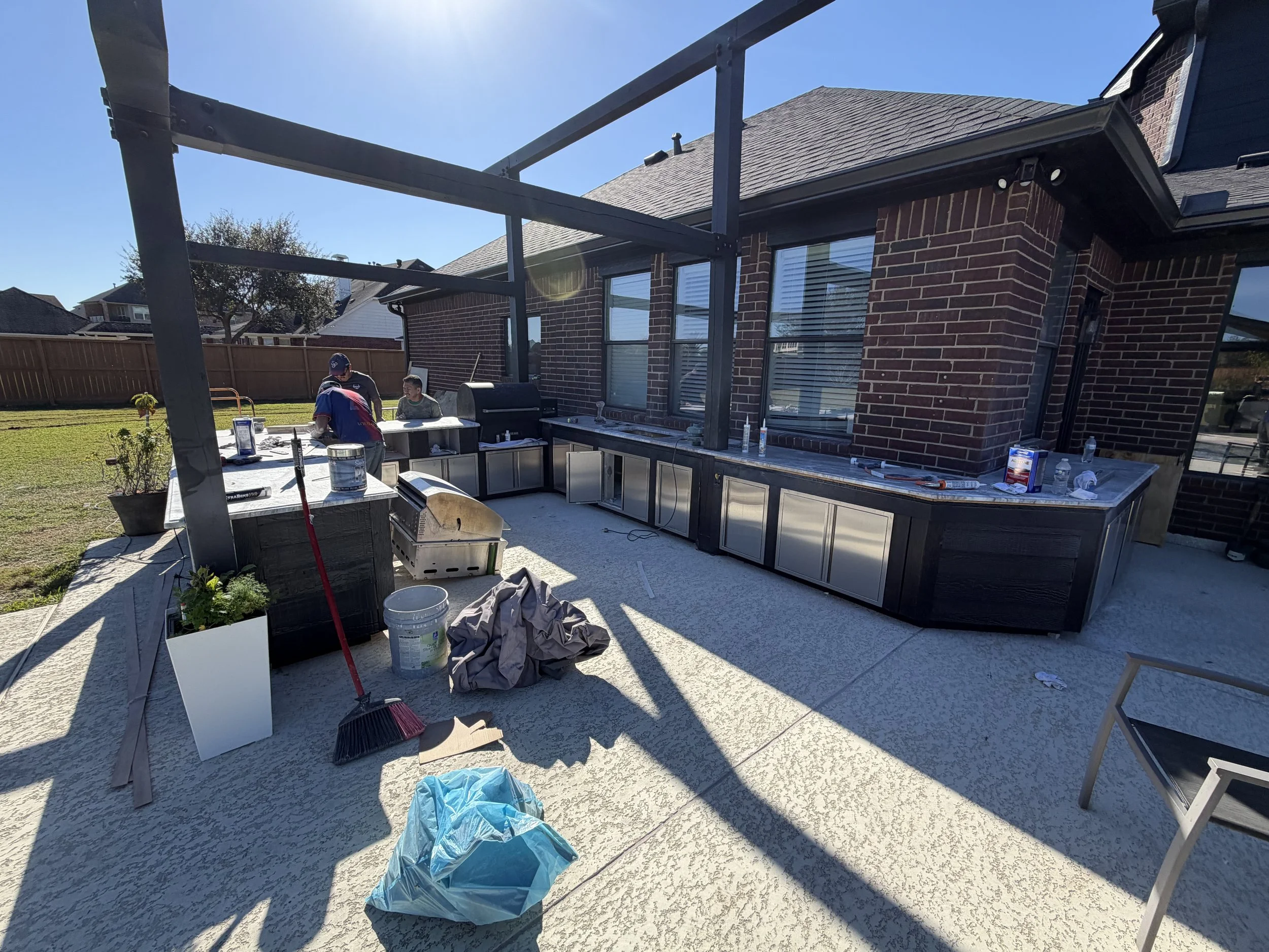 Backyard patio area under construction with outdoor kitchen, construction tools, and two workers, in sunny weather.