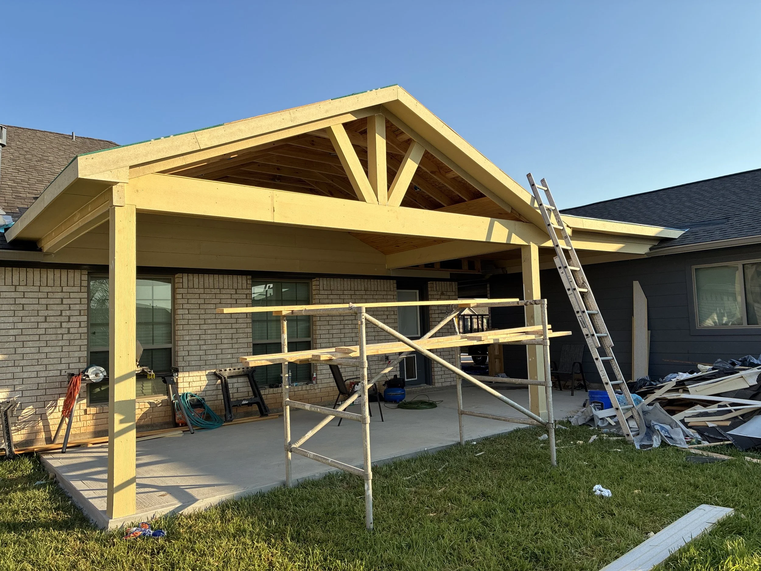 Construction of a new porch or patio extension on a brick house with a wooden roof structure, scaffolding, and construction materials around, in a backyard during daytime.