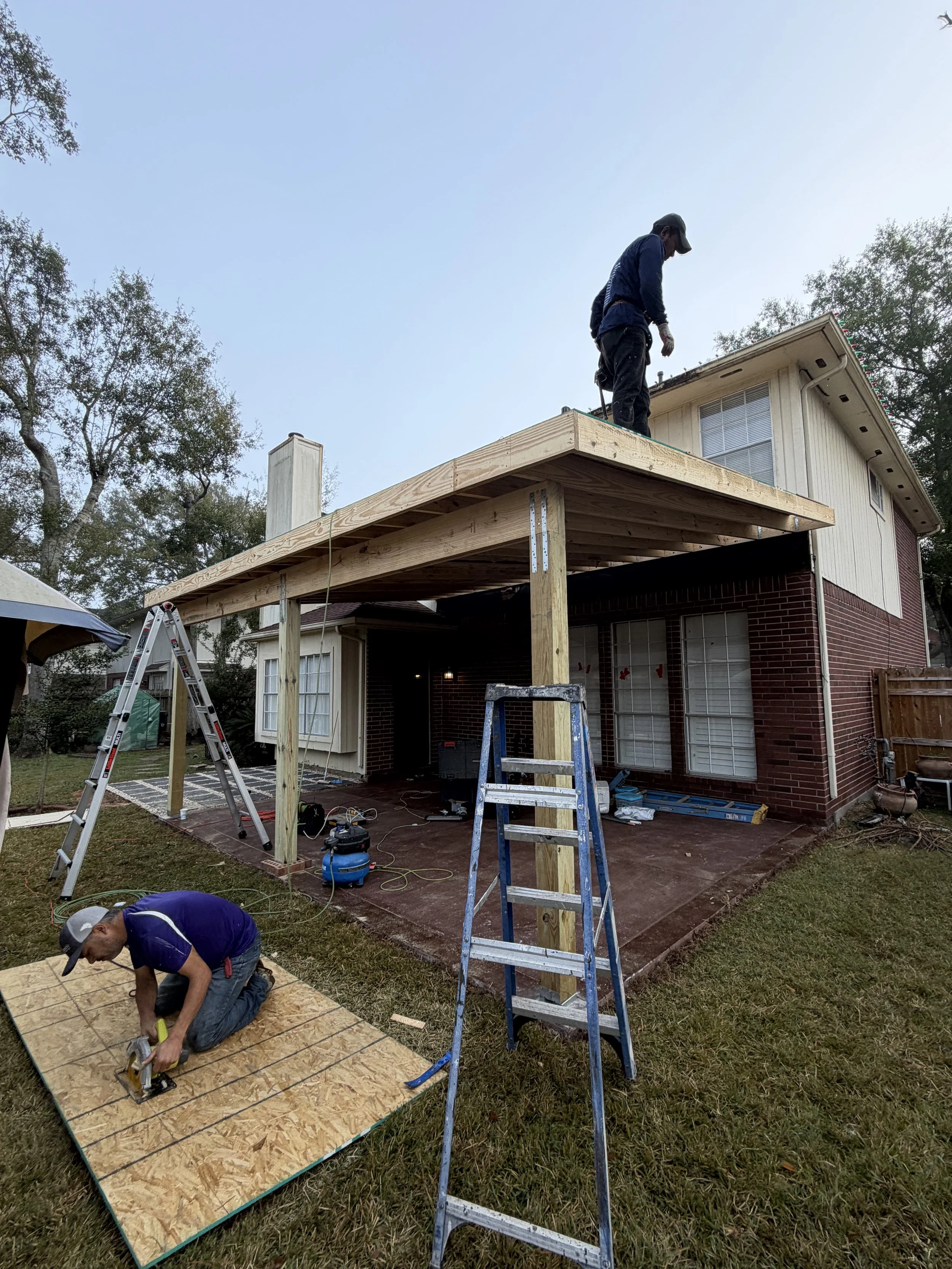 Two workers building a large wooden deck or porch extension on the back of a house. One worker is kneeling on a sheet of plywood on the ground, cutting with a saw, while the other is standing on the new deck structure.