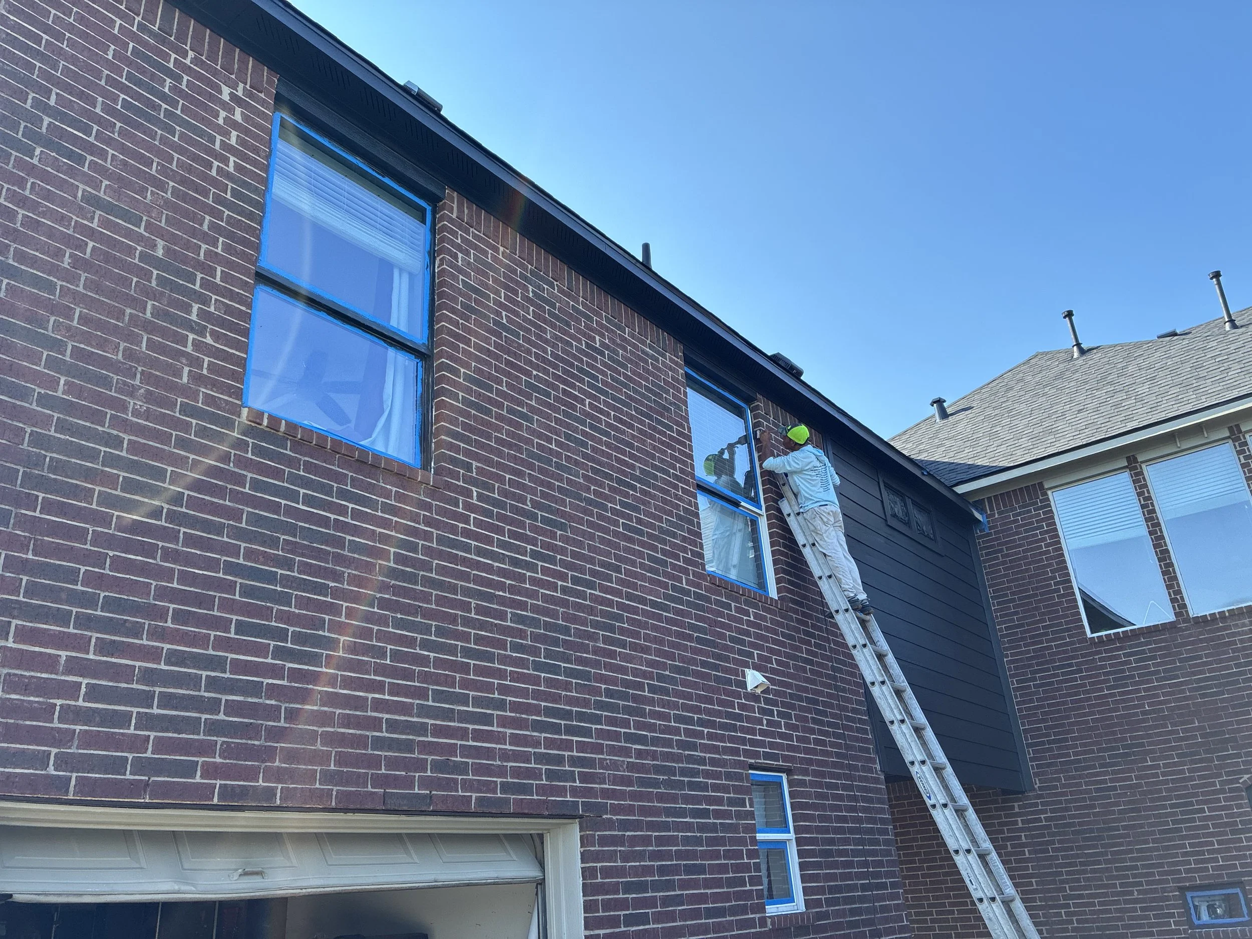 A person in white clothing and a yellow helmet is working on a second-floor window of a brick house, standing on a tall extension ladder.